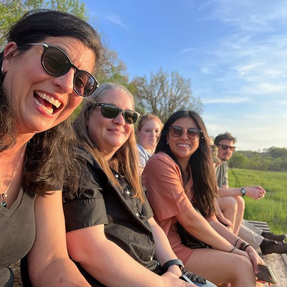 A group of people sitting outside, smiling at the camera. Sunny day with green field in background.