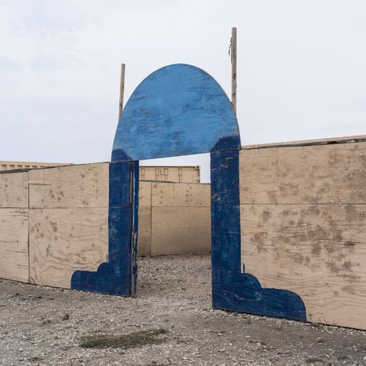 Blue painted archway on plywood structure, in a gravel lot under an overcast sky.