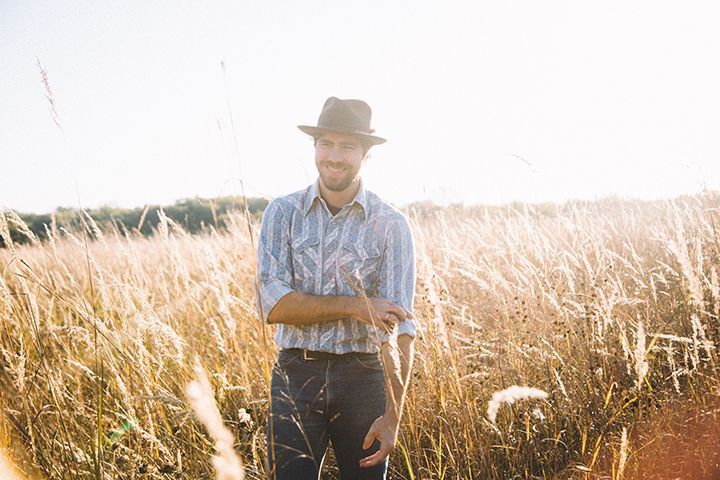Man in a hat standing in tall, golden grass field; sunlight, looking down, rolling up his sleeve.