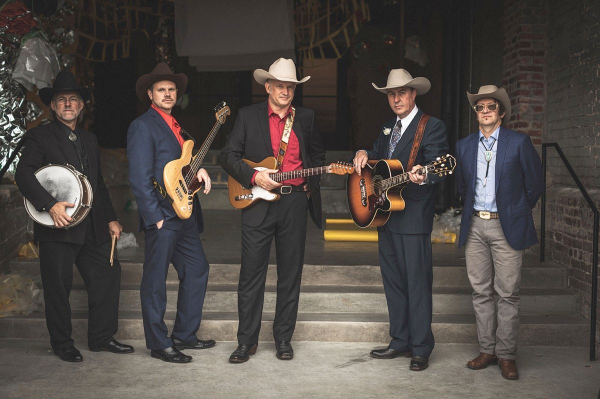 Five men in cowboy hats and suits with musical instruments, posing outside.