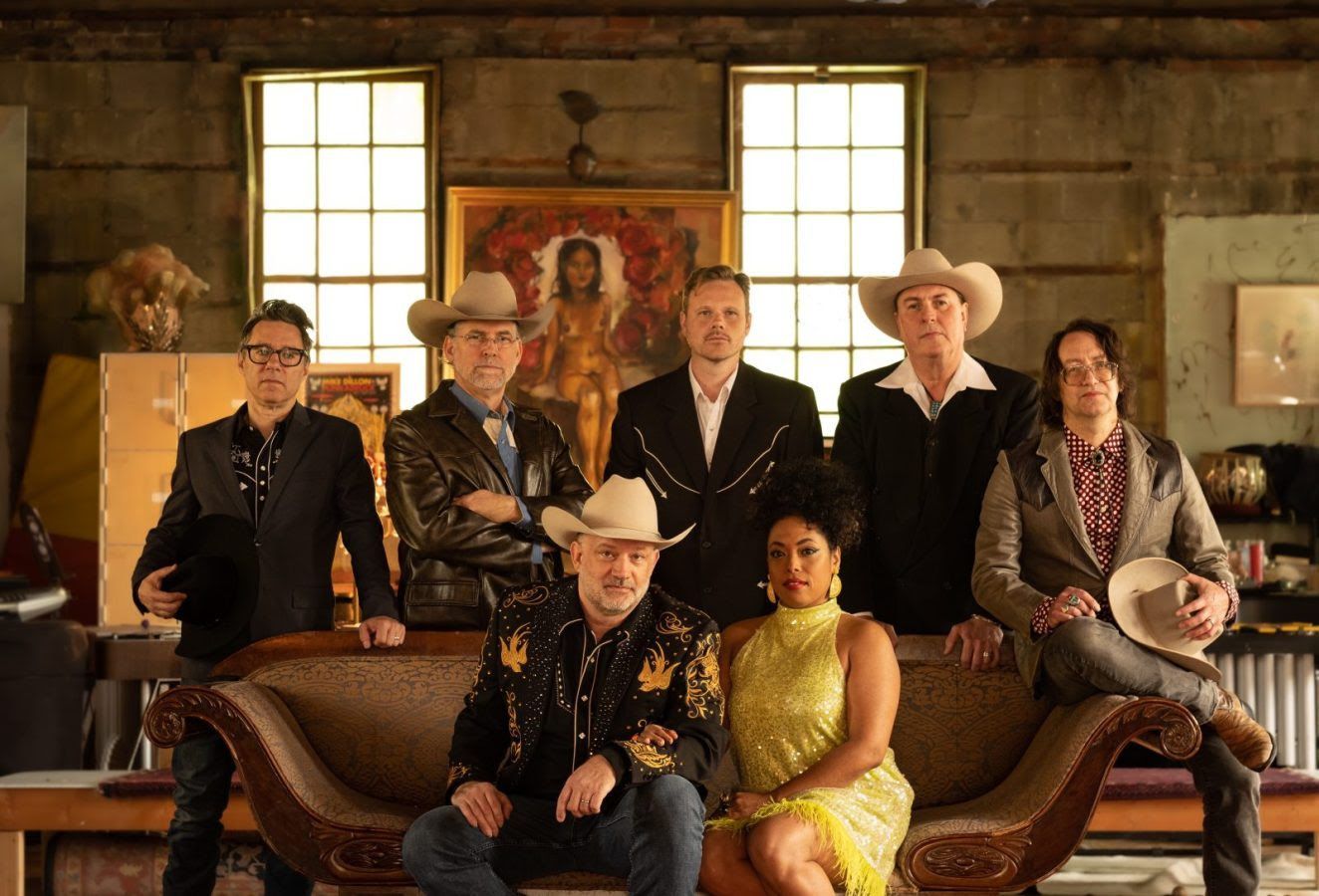 Band of seven people pose indoors; some wear cowboy hats, a woman wears yellow, antique couch.