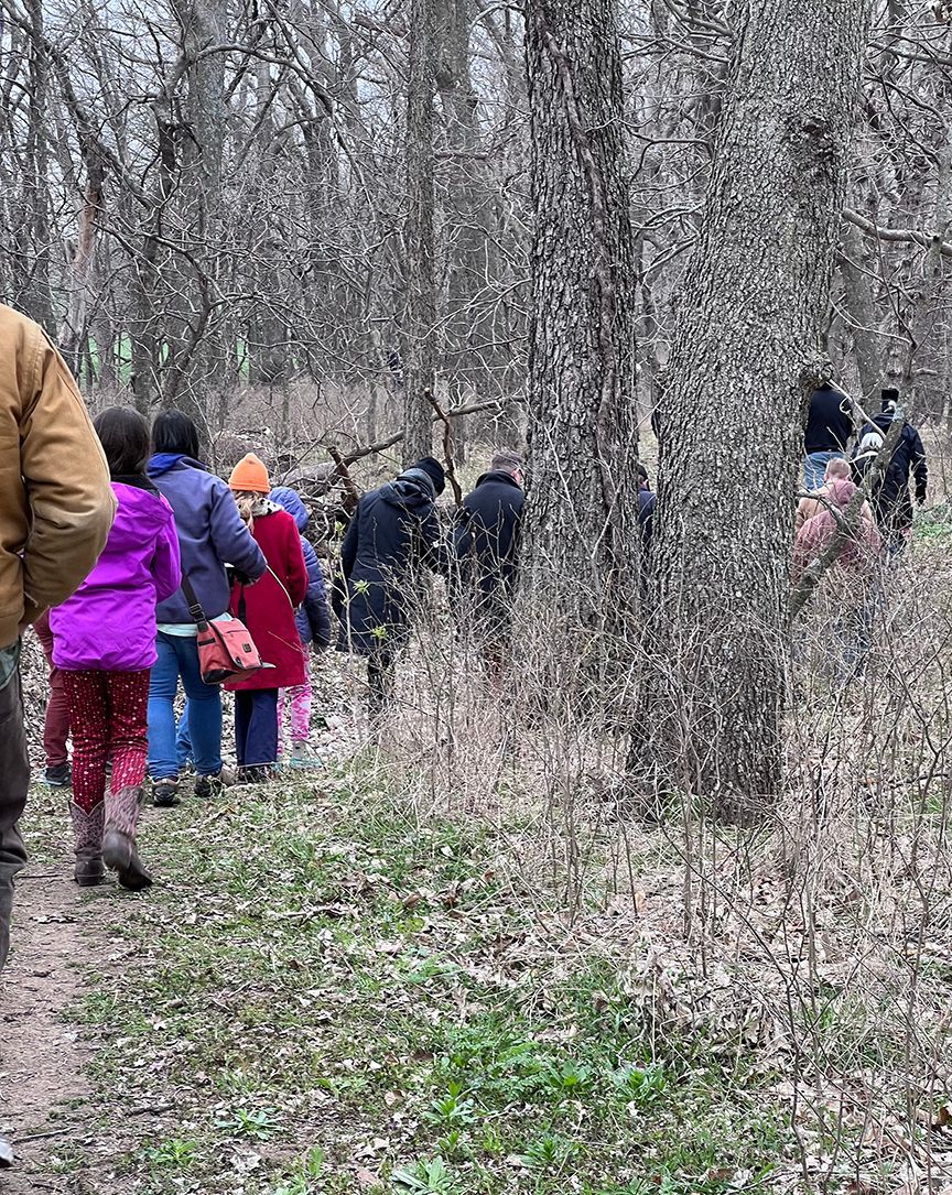 People walking single file through a wooded area. They wear coats and backpacks. Bare trees surround them.