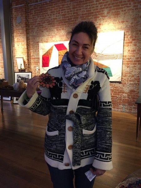 A woman wearing a sweater and scarf is holding a donut in front of a brick wall