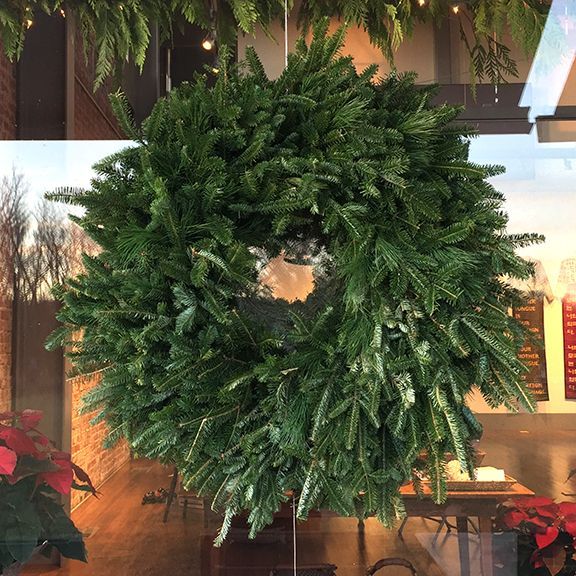 A large, green Christmas wreath hanging in a window, with a partial view of a room and poinsettias.