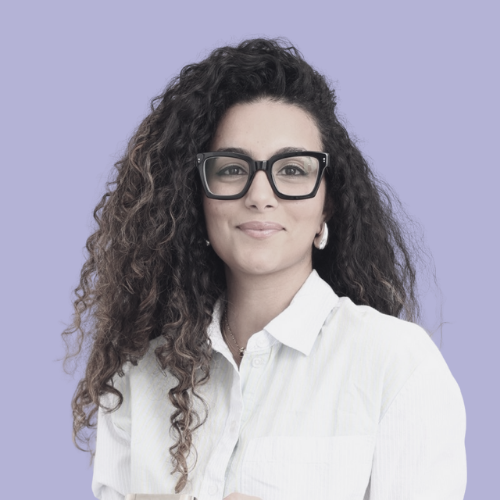 Oumaima Talouka. She has curly hair and glasses.  She is smiling, wearing a white shirt against a purple background.