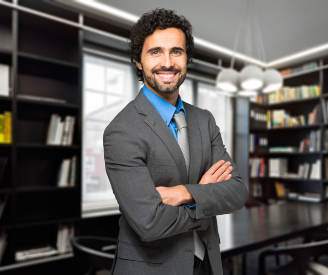 Man in a gray suit with arms crossed, smiling in an office with bookshelves.