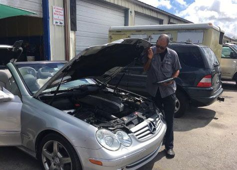 Mechanic inspecting car engine with open hood, holding clipboard, sunny outdoor shop setting.