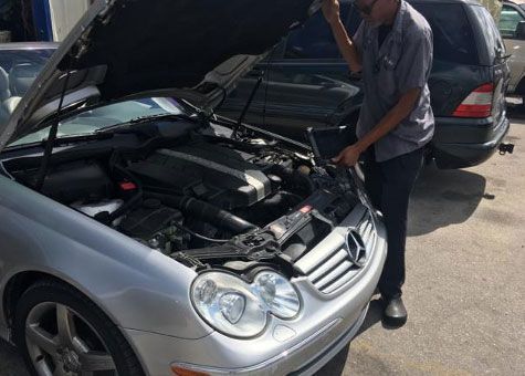 Mechanic checking car engine with hood open; silver Mercedes in sunlight.