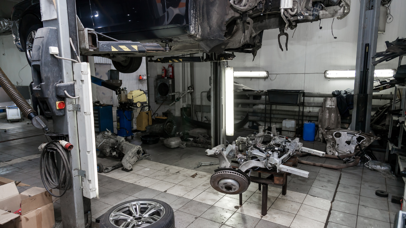 Car being worked on in a cluttered auto repair shop. Engines and parts scattered around.