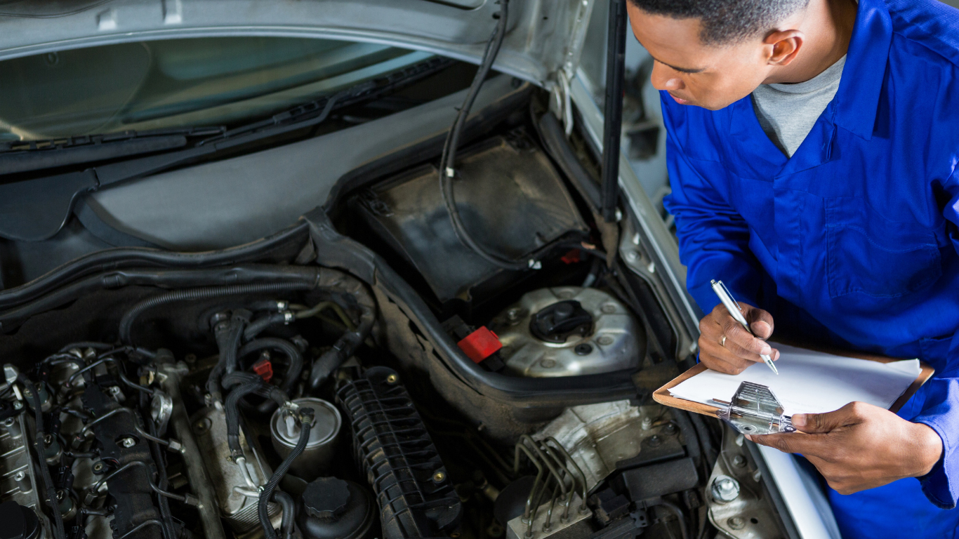 Mechanic in blue jumpsuit inspecting car engine, taking notes on clipboard.
