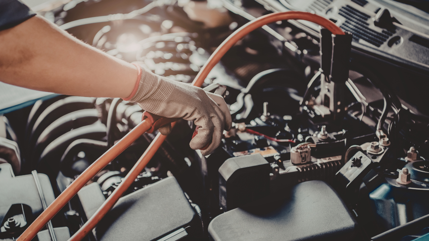 Gloved hand connecting jumper cable to a car battery in an engine bay.