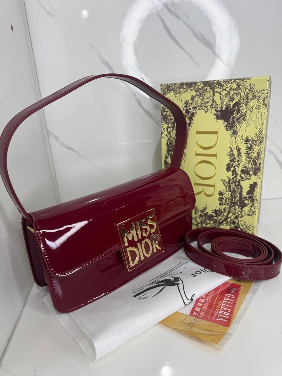Bolso Miss Dior color burdeos con caja y correa sobre superficie blanca.
