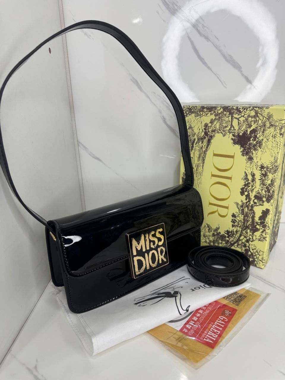 Bolso de hombro "Miss Dior" de charol negro con accesorios, sobre superficie blanca.