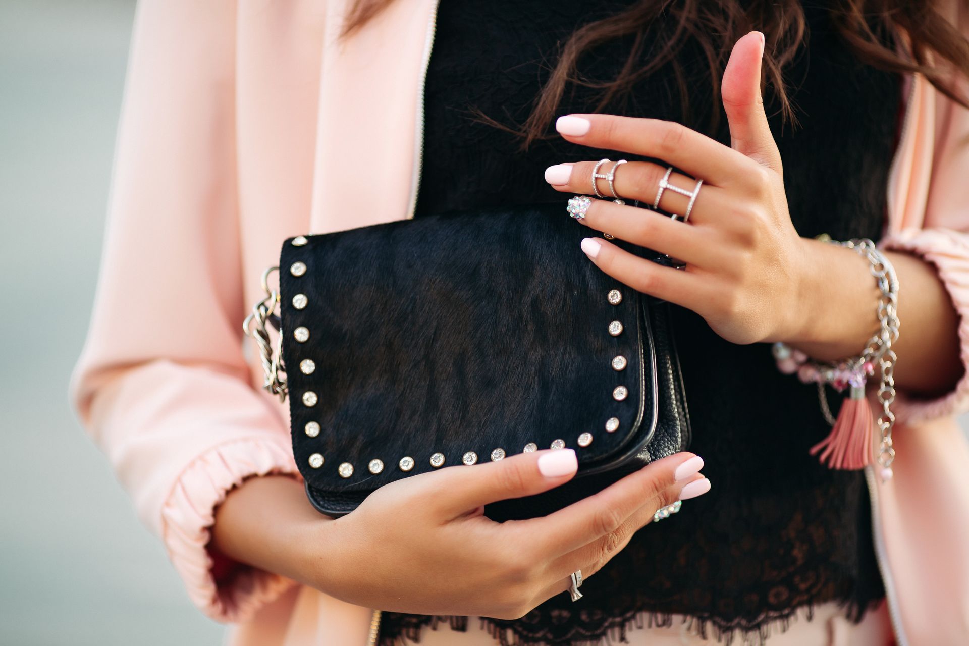 Mujer sosteniendo un bolso negro con tachuelas, luciendo anillos, pulseras, blazer rosa y top de encaje.