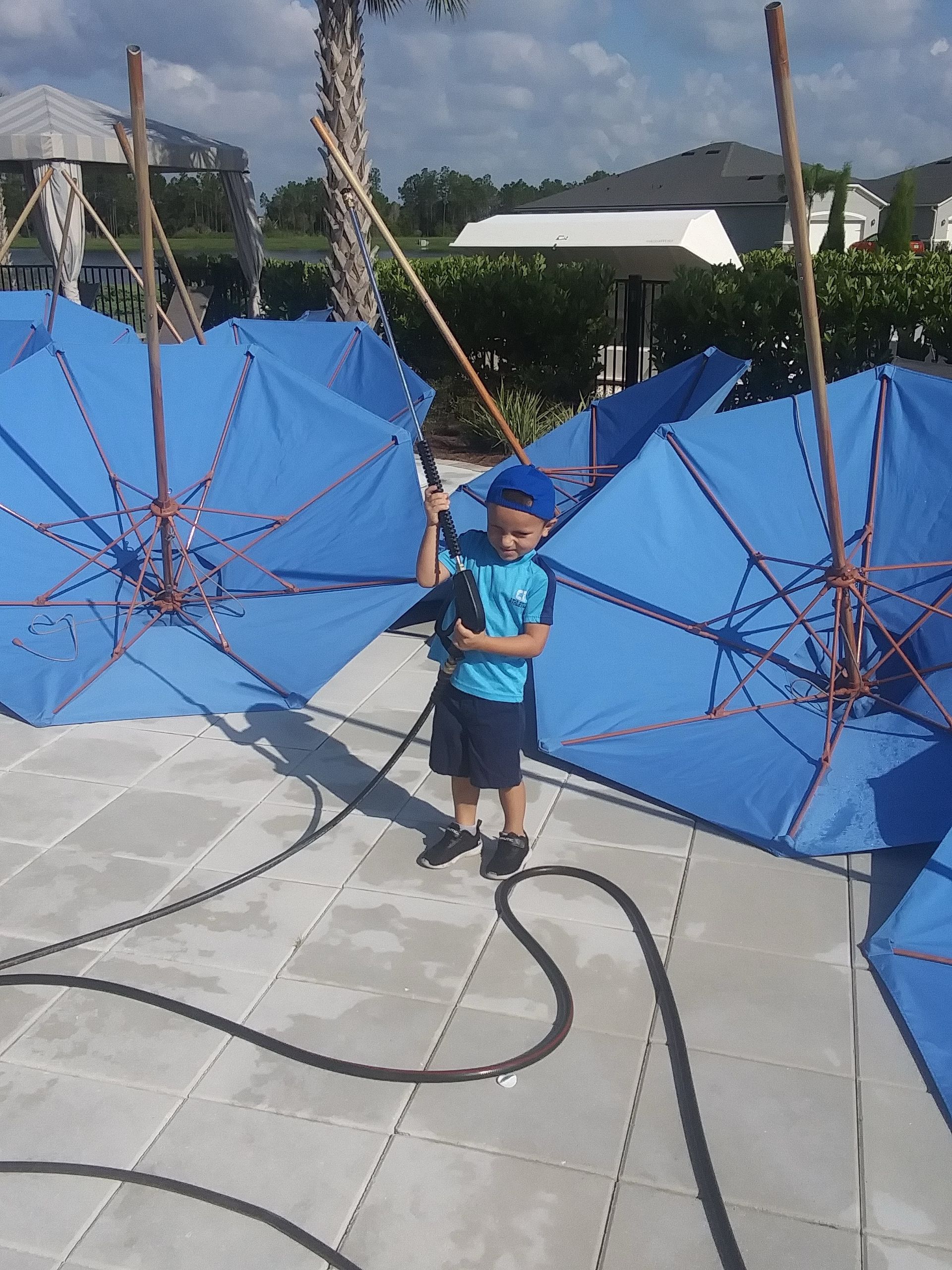 Boy in blue shirt and hat standing among blue umbrellas on a sunny patio.