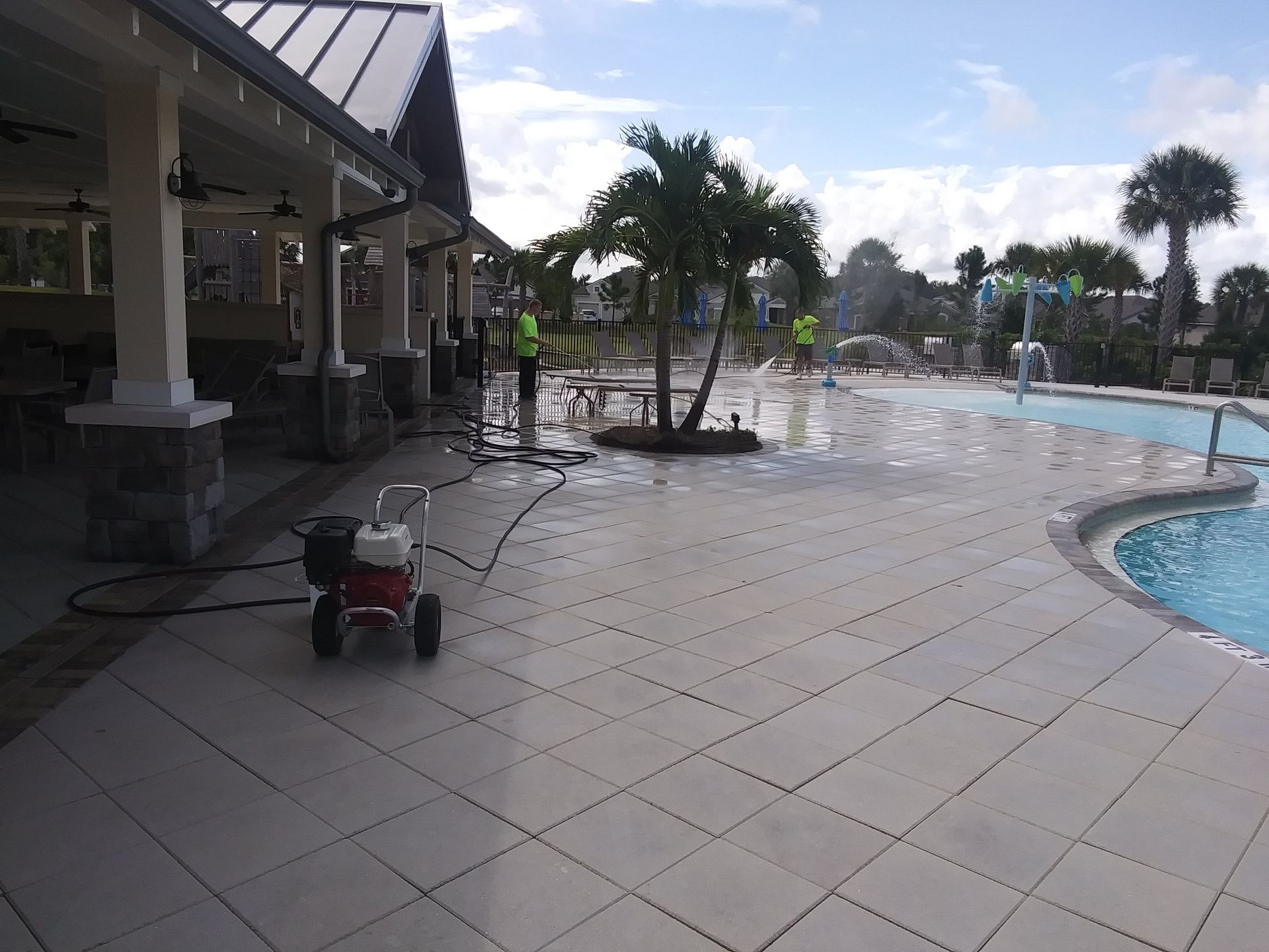 Workers pressure washing a tiled patio next to a pool and a covered outdoor area.