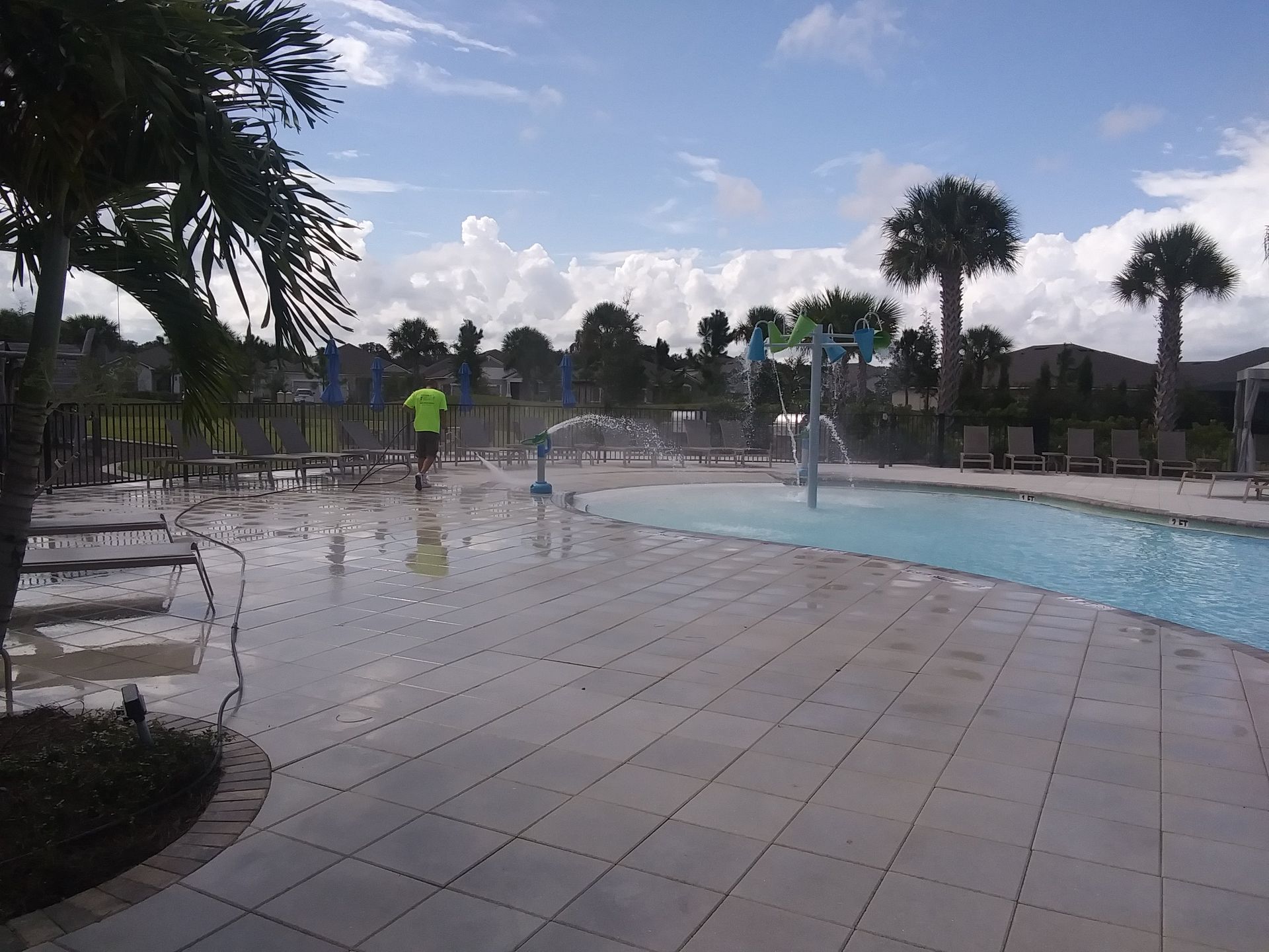 A splash pad with water features and a pool on a sunny day, with palm trees and a person in the distance.