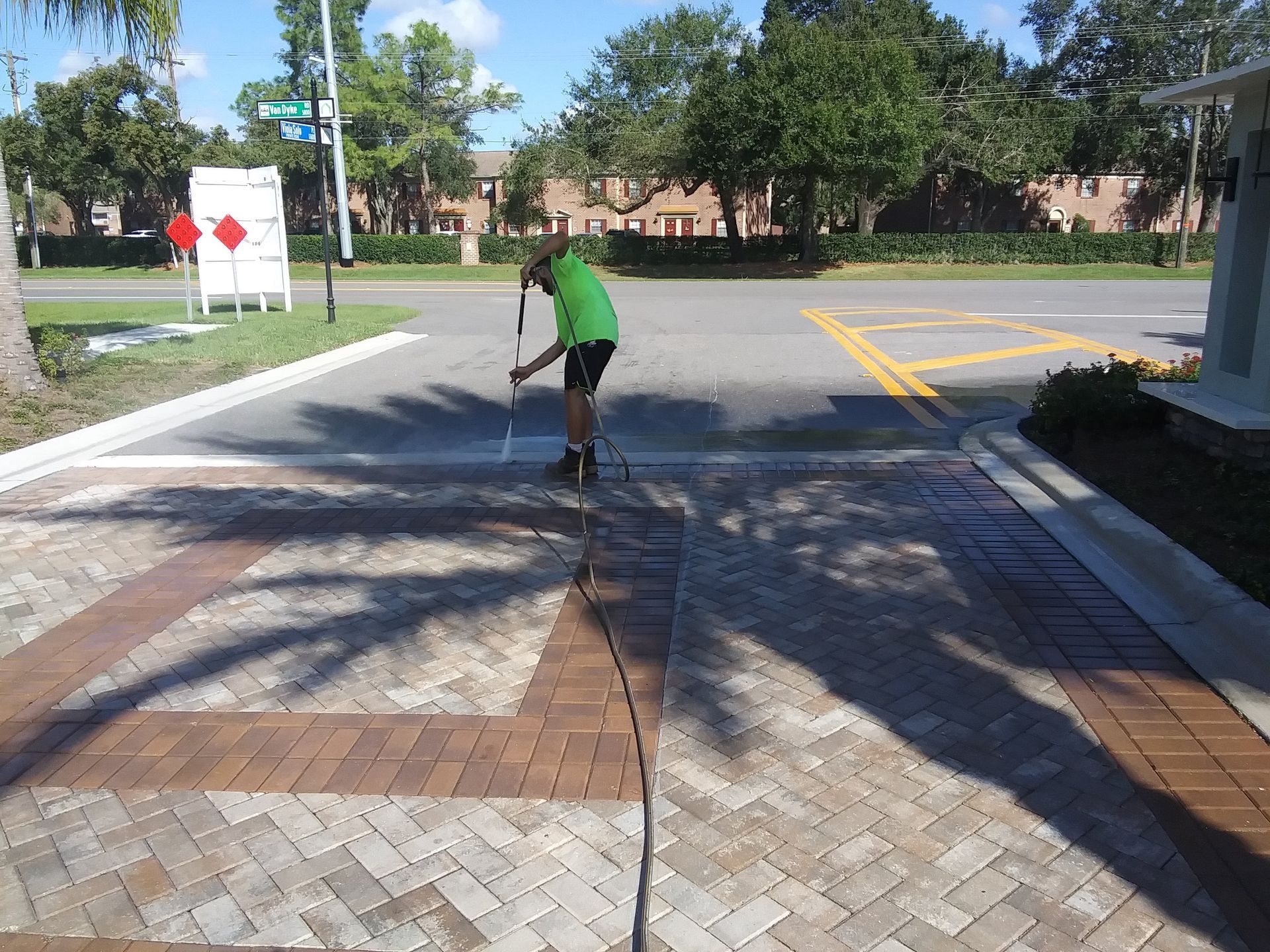 Person in green shirt cleaning brick sidewalk with pressure washer near a street.