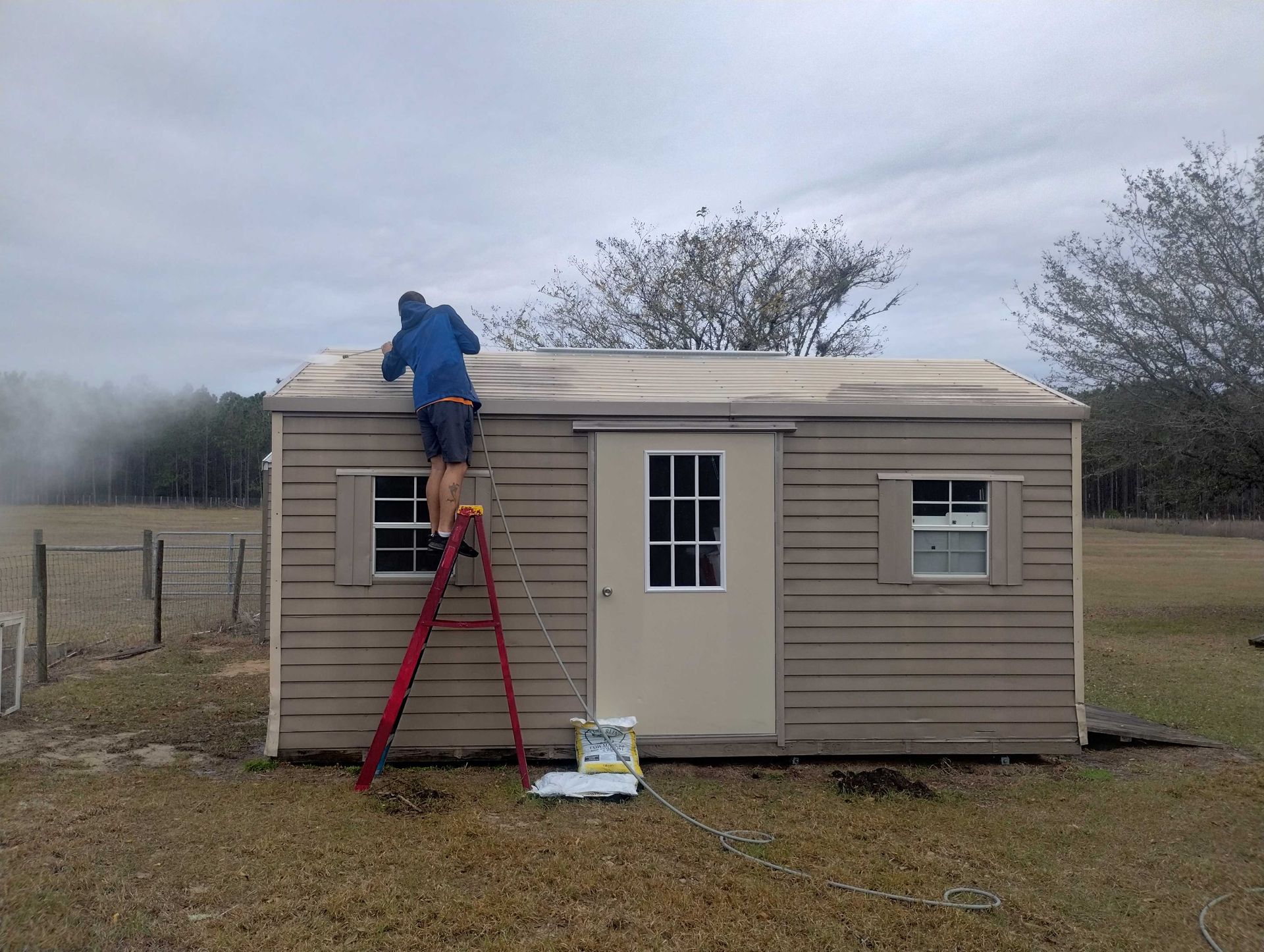 Man on a ladder repairing shed roof. Brown shed in a field on an overcast day.