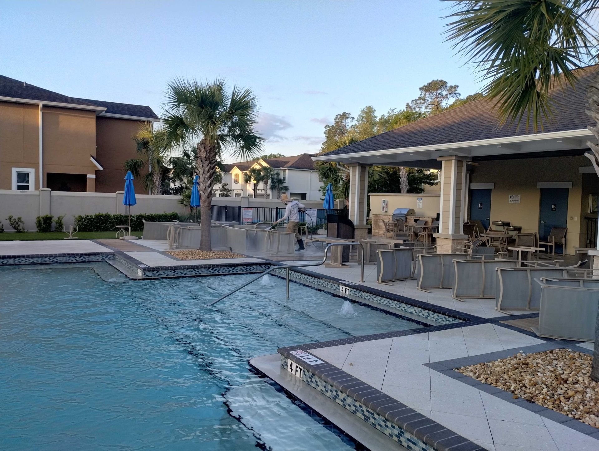 Swimming pool with fountains, lounge area, and buildings under a blue sky.