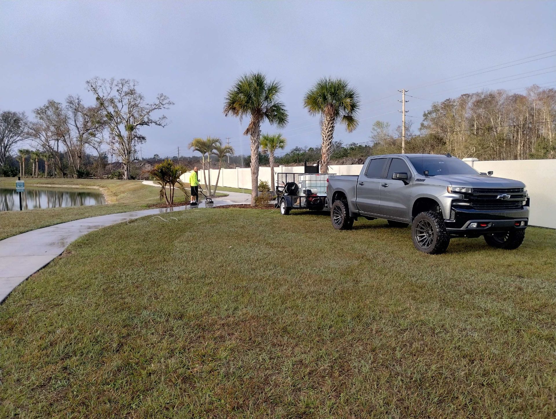 Gray truck with a trailer on a grassy lawn near a pond, a person in a yellow vest nearby.