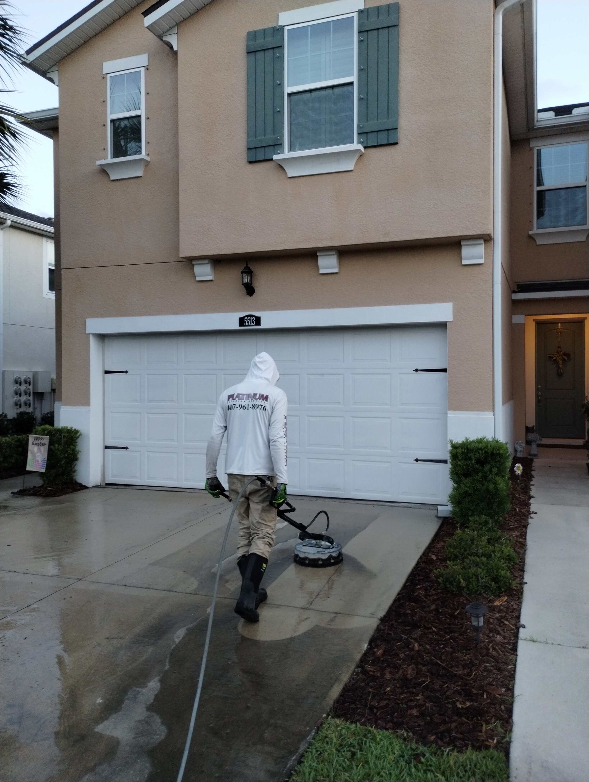 Person in protective gear power washing a driveway in front of a beige two-story house with a white garage door.