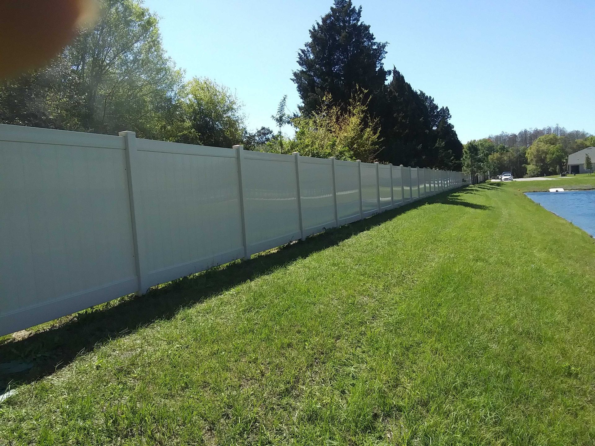 White fence along a grassy bank next to a pond, trees in the background on a sunny day.