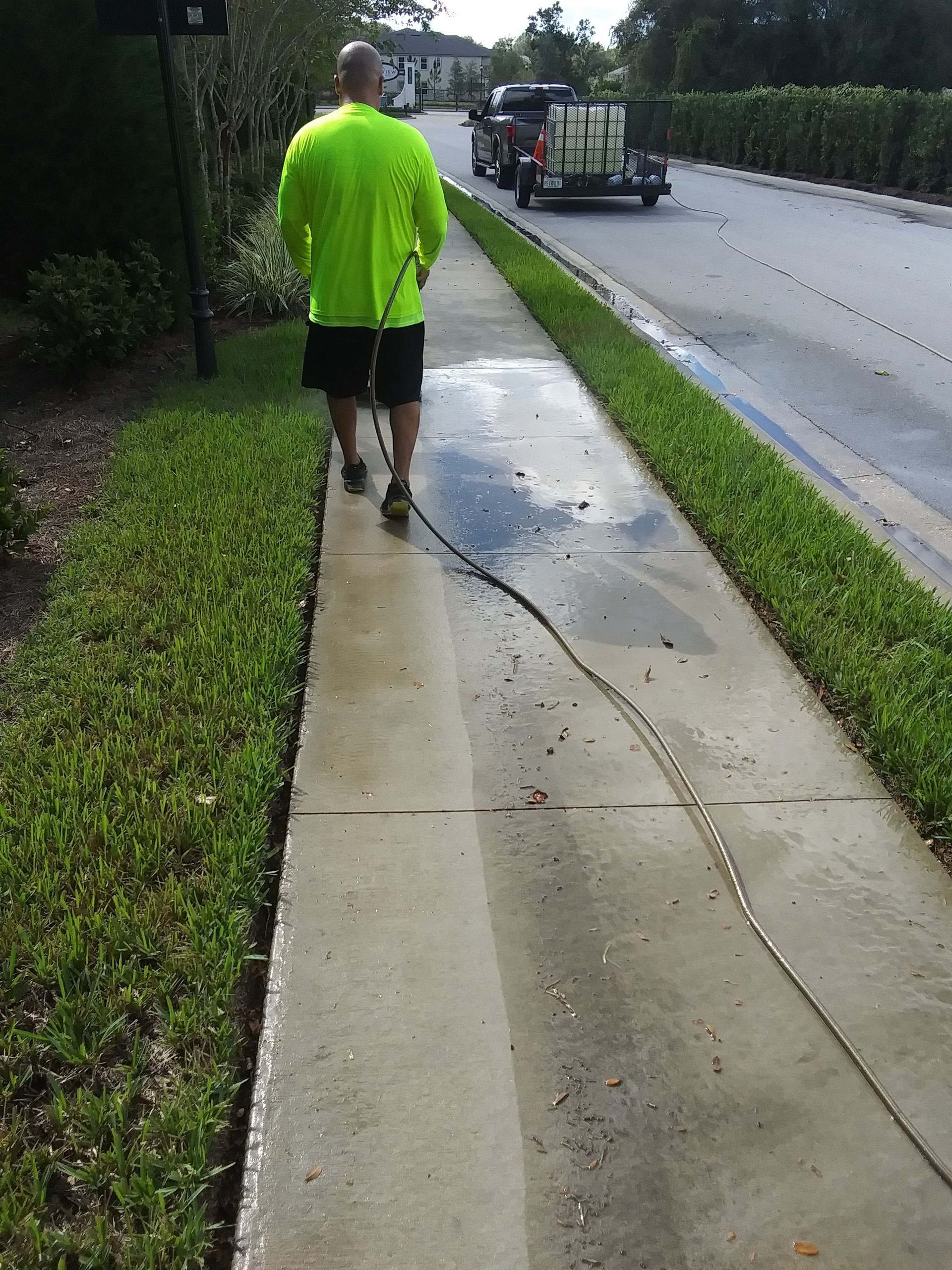 Man pressure washing a sidewalk next to grass and a road, with a vehicle in the background.