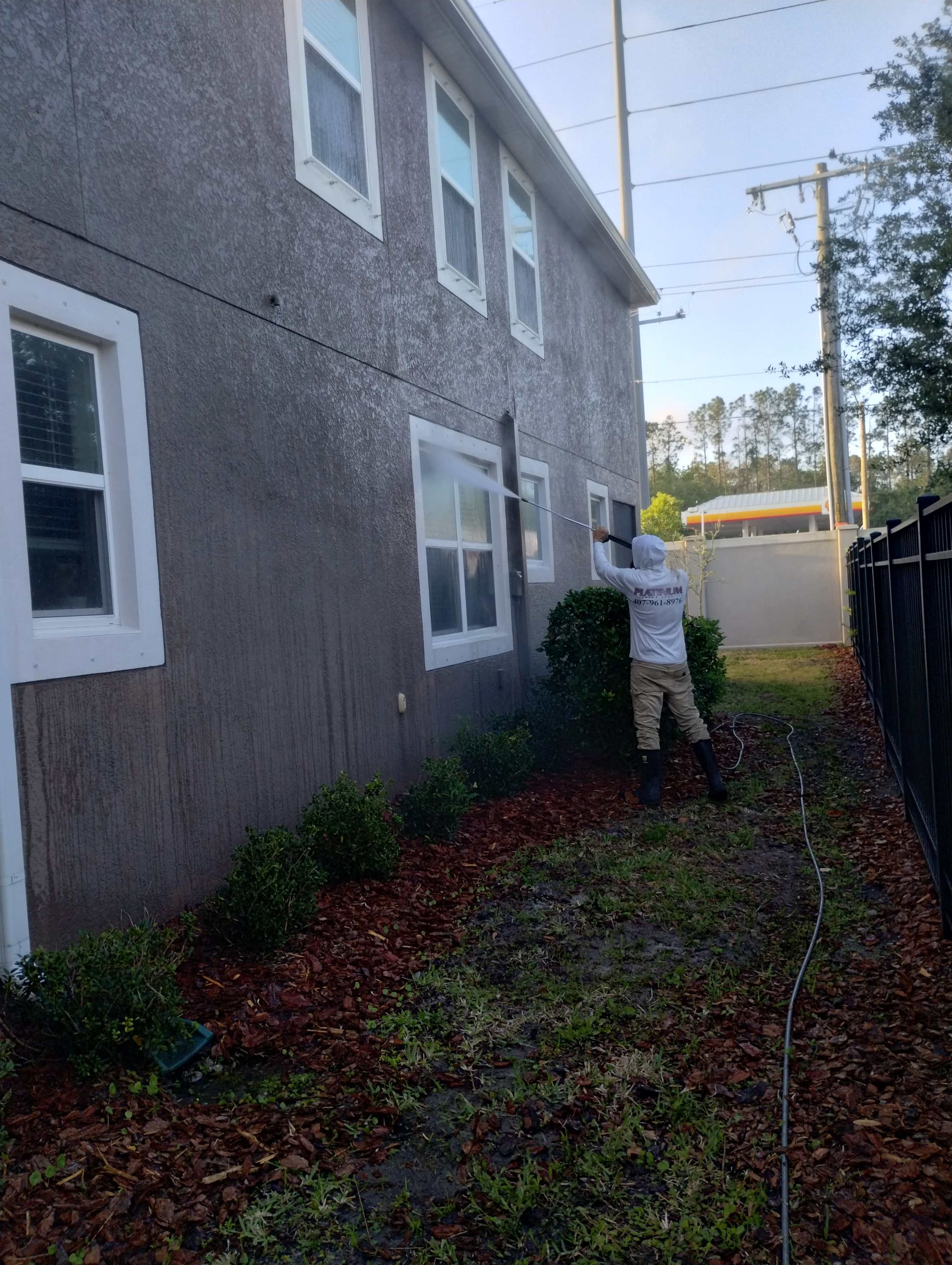Person power washing a two-story house with gray stucco walls, green yard, and black fence.