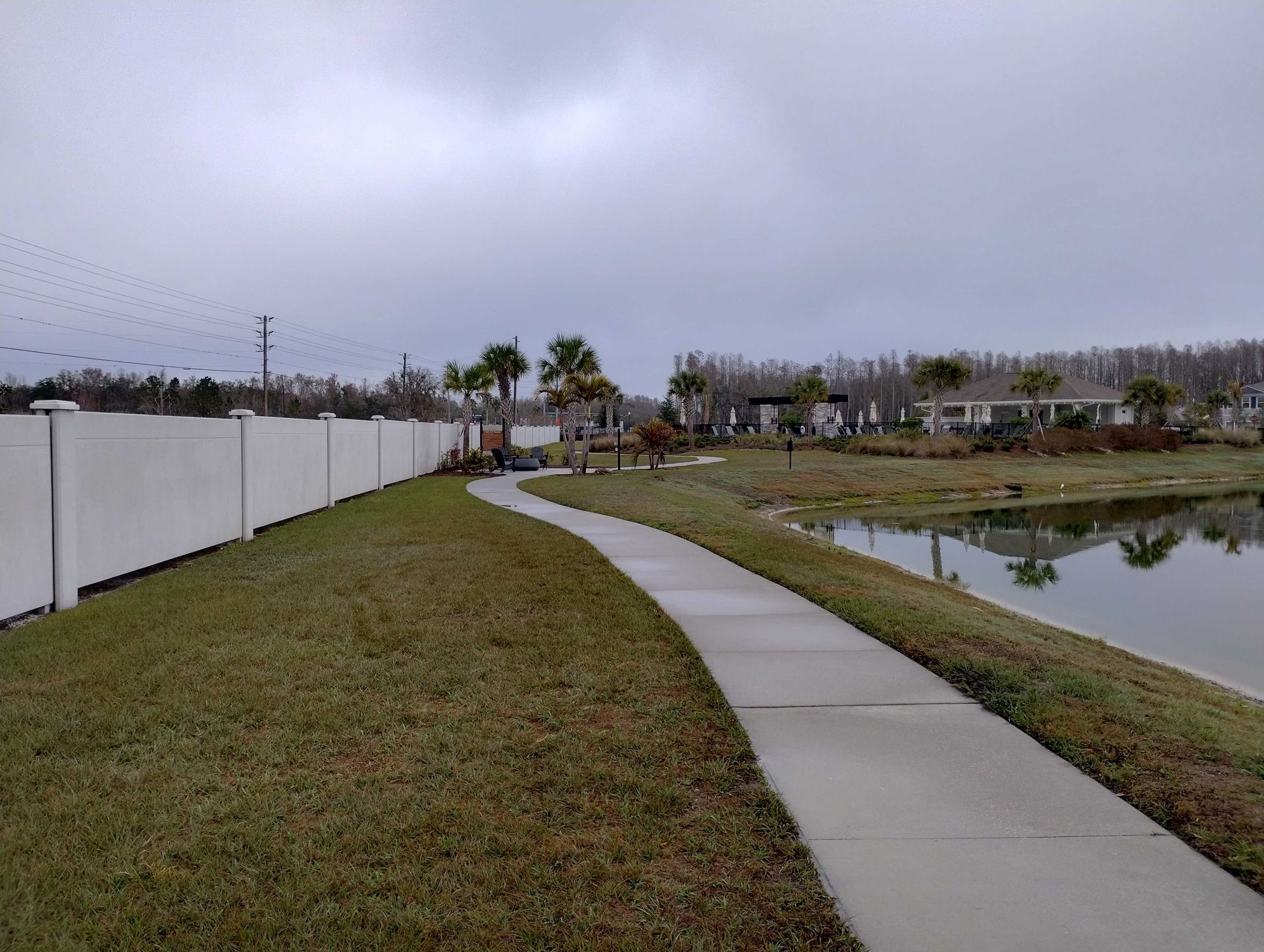 A paved pathway curves along a pond, bordered by a white fence and grass, under a cloudy sky.