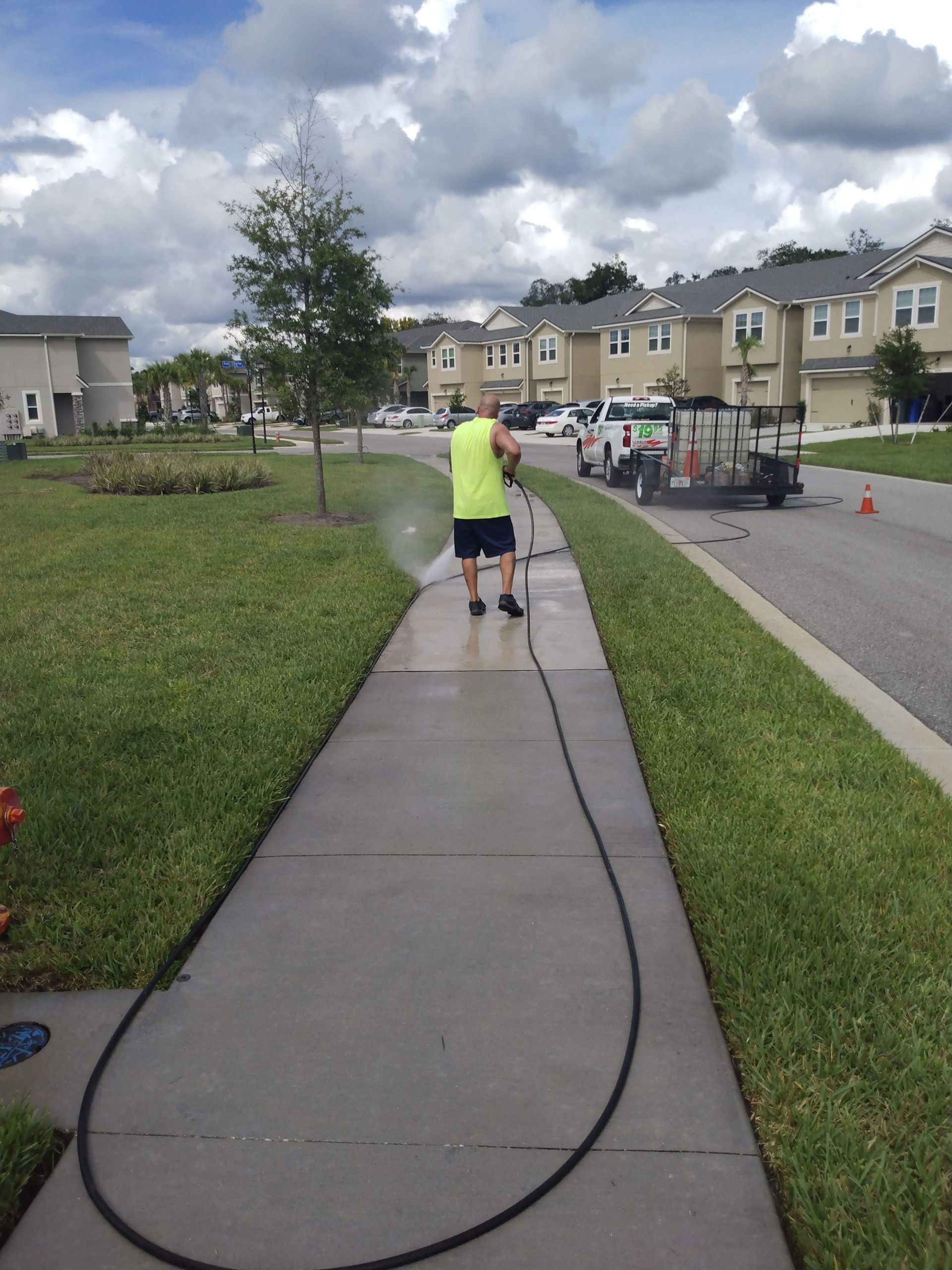 Man pressure washing a sidewalk in a residential neighborhood with a green lawn on either side.