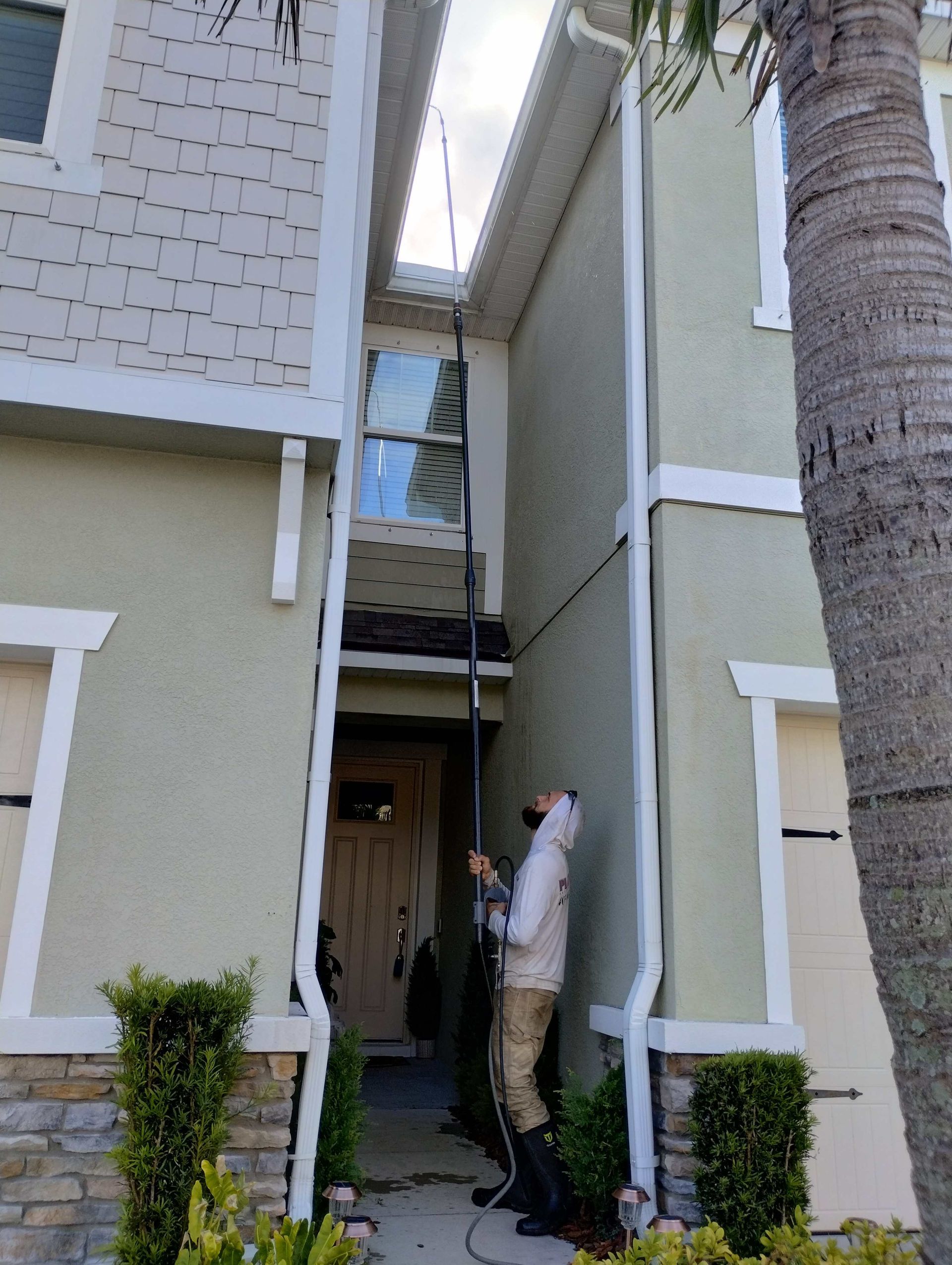 Person using a pole to clean gutters on a two-story house, standing between two buildings.