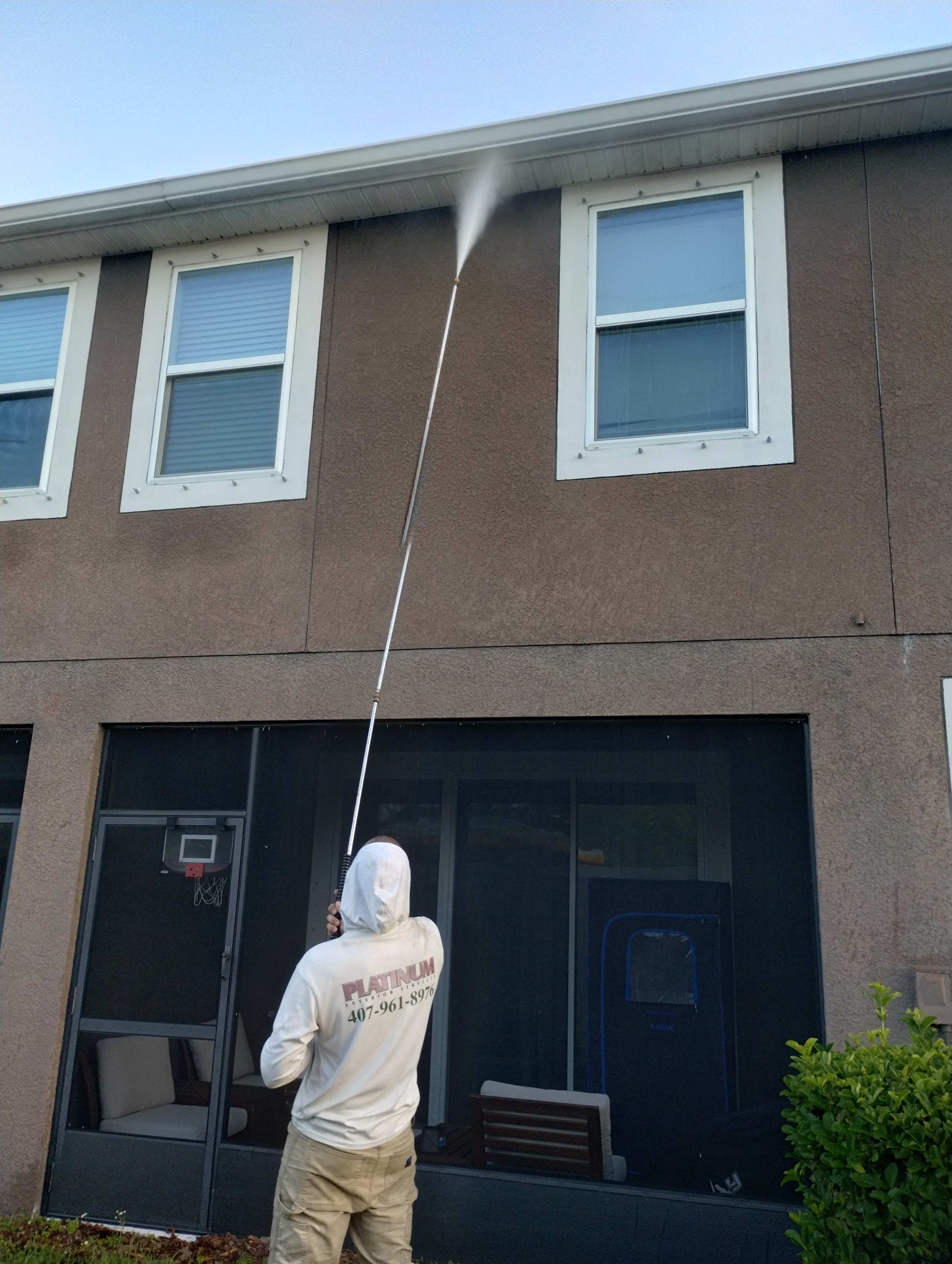 Man pressure washing the side of a two-story building with white trim and windows, using an extended wand.