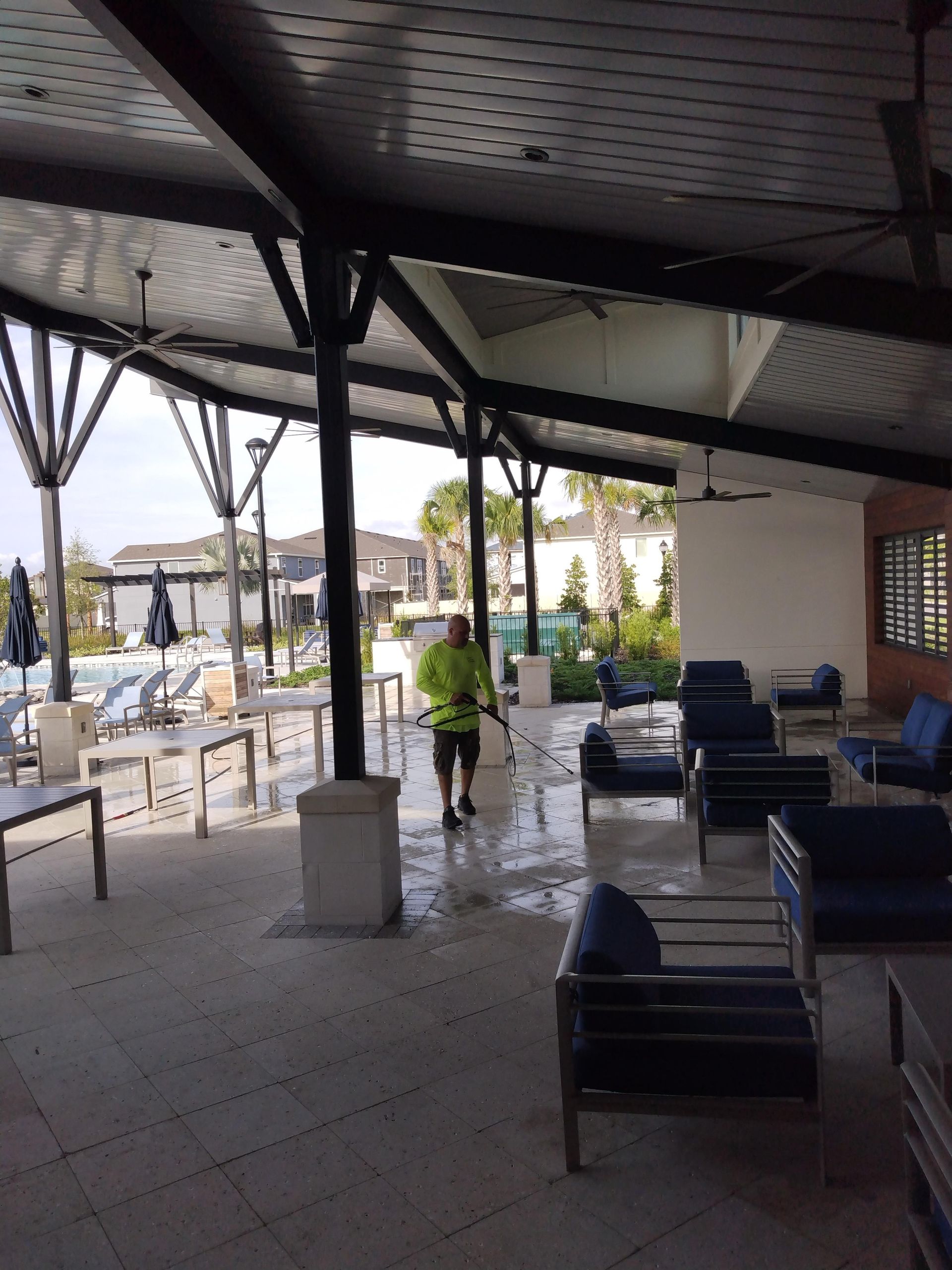 A person wearing a yellow vest power washes an outdoor patio with seating near a pool.