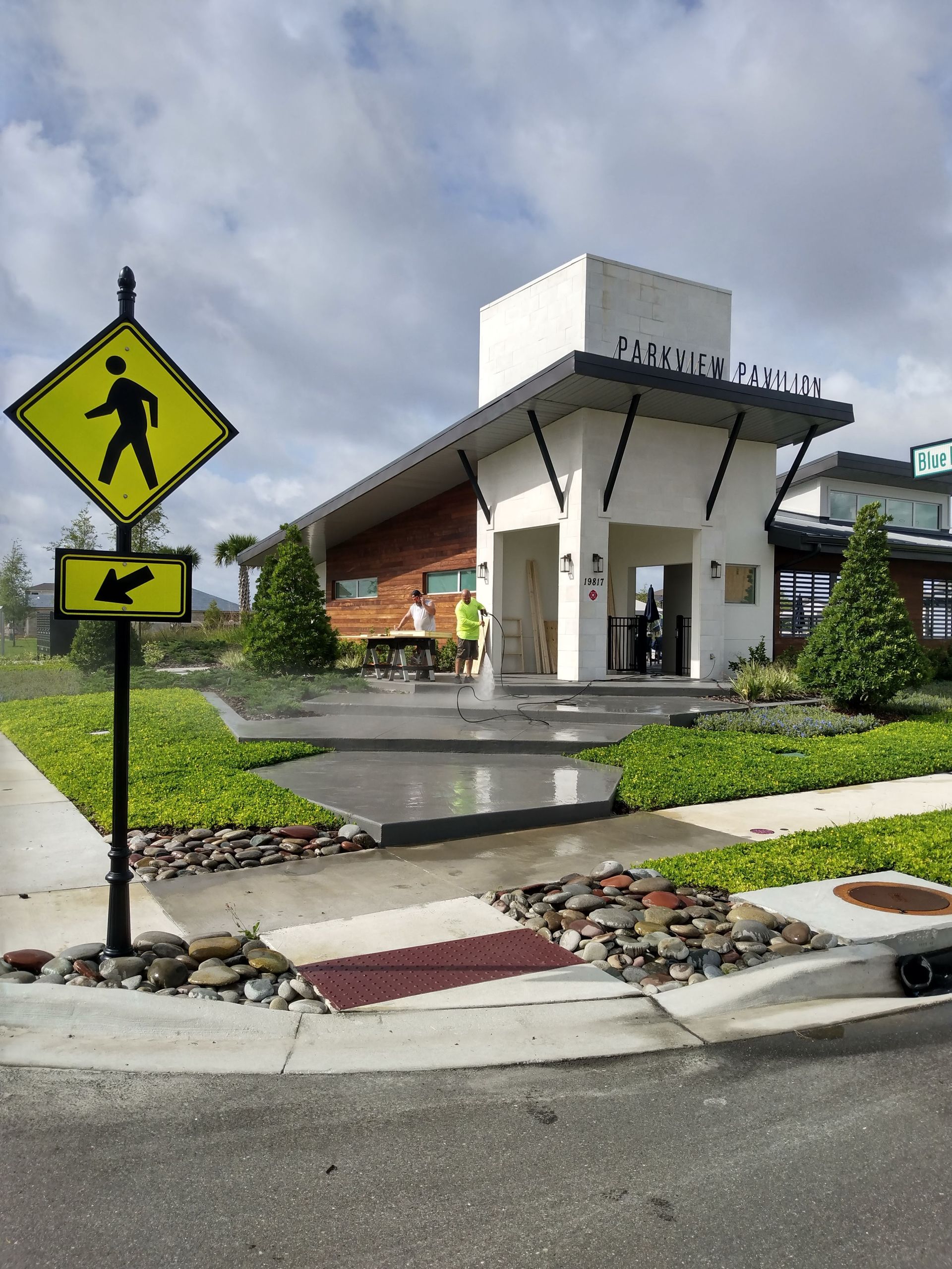 Yellow pedestrian crossing sign at a building entrance, landscaping and a cloudy sky in the background.