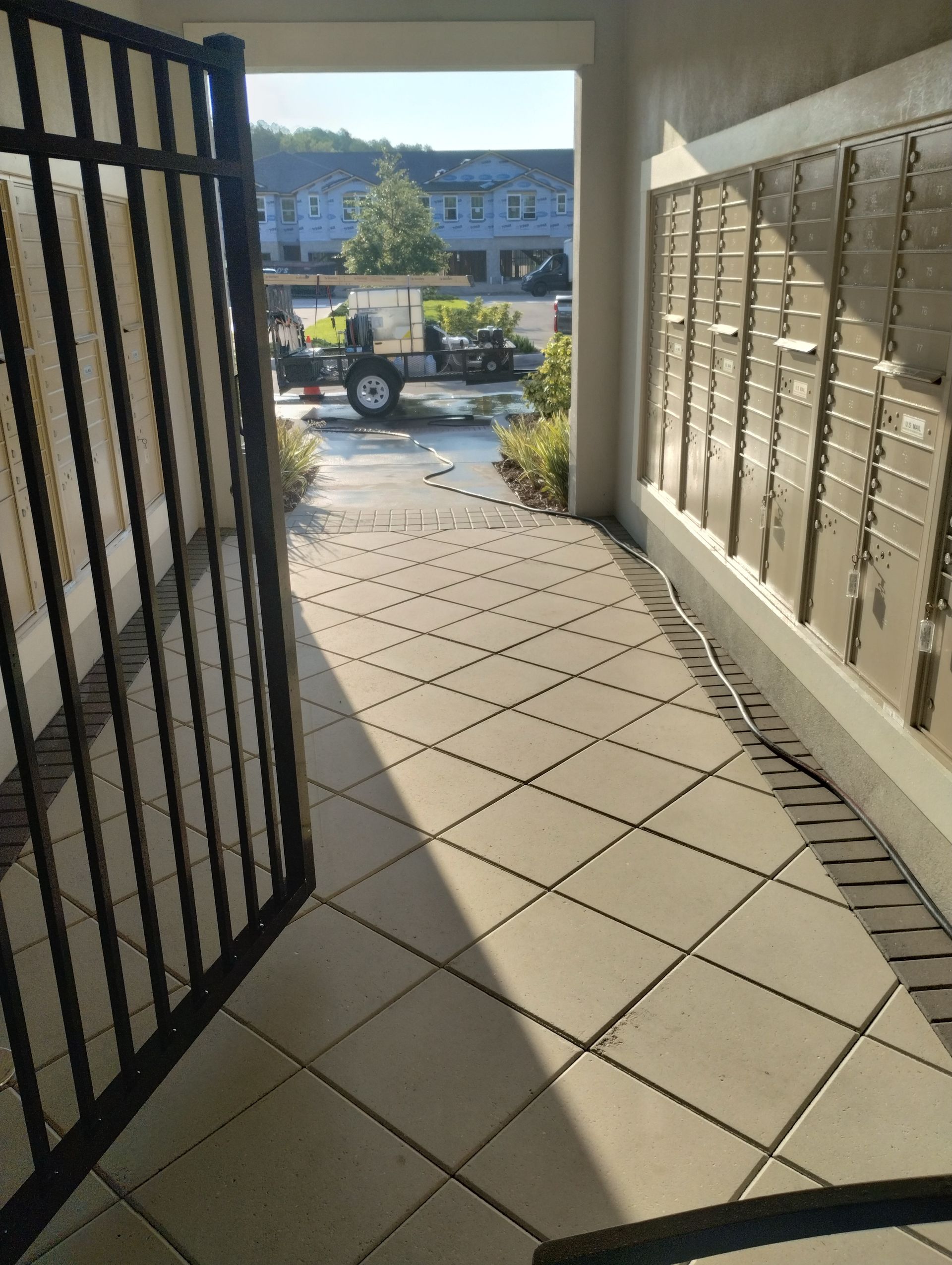 Entrance to mailboxes with a gate, tiled walkway, and a view of a vehicle and buildings.