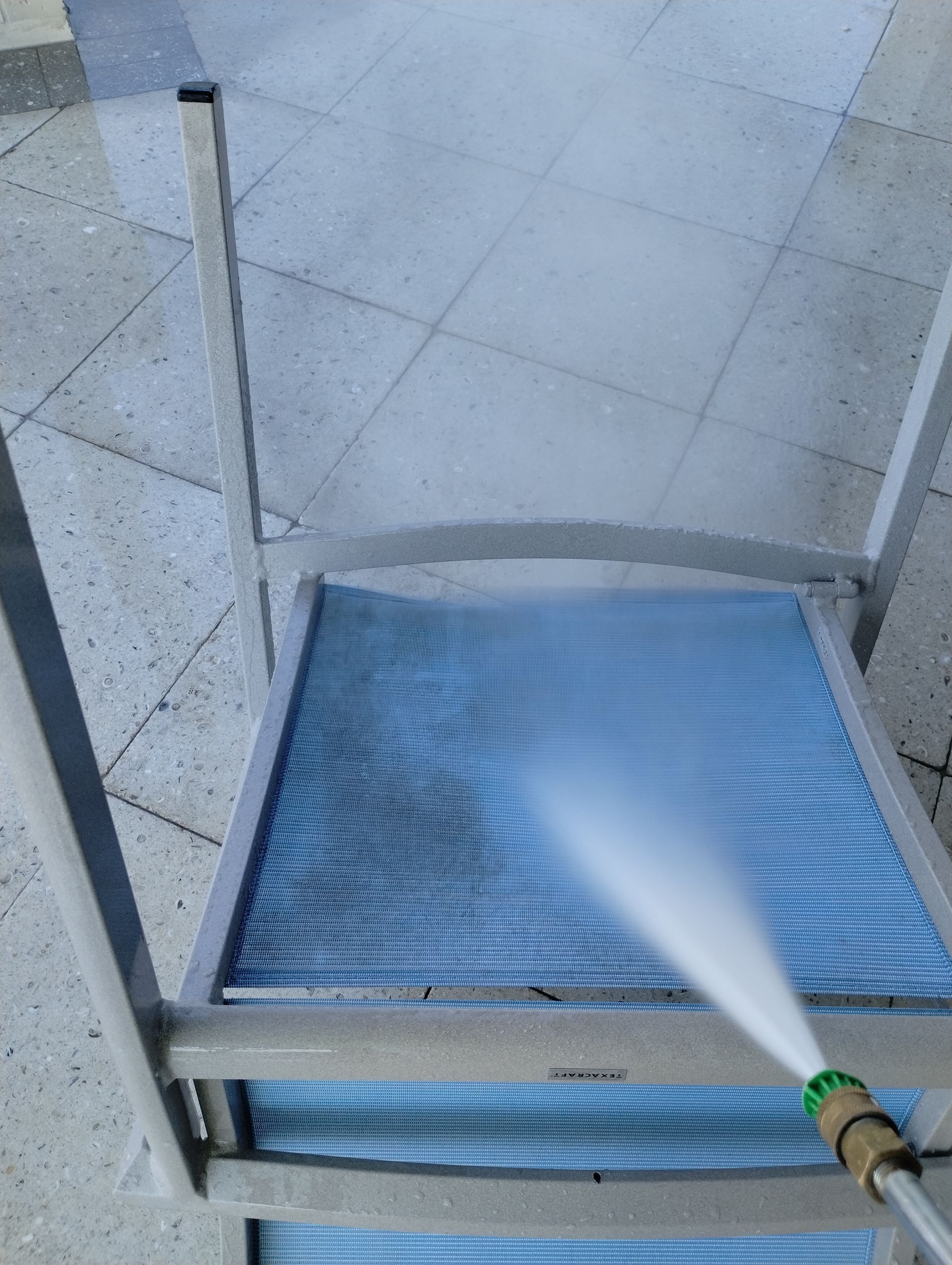 Person spraying water on a blue mesh chair with a hose outside.