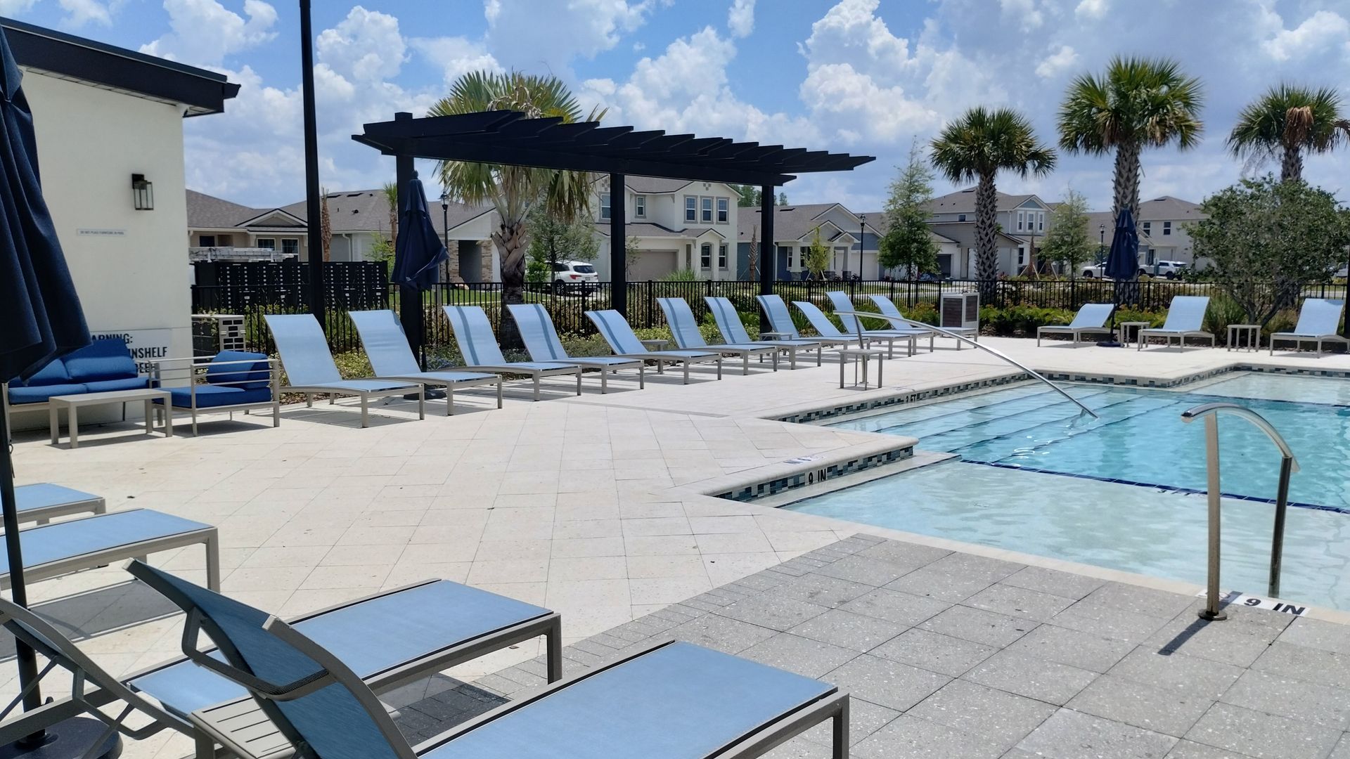 Poolside scene with lounge chairs, a pergola, and palm trees under a bright blue sky.