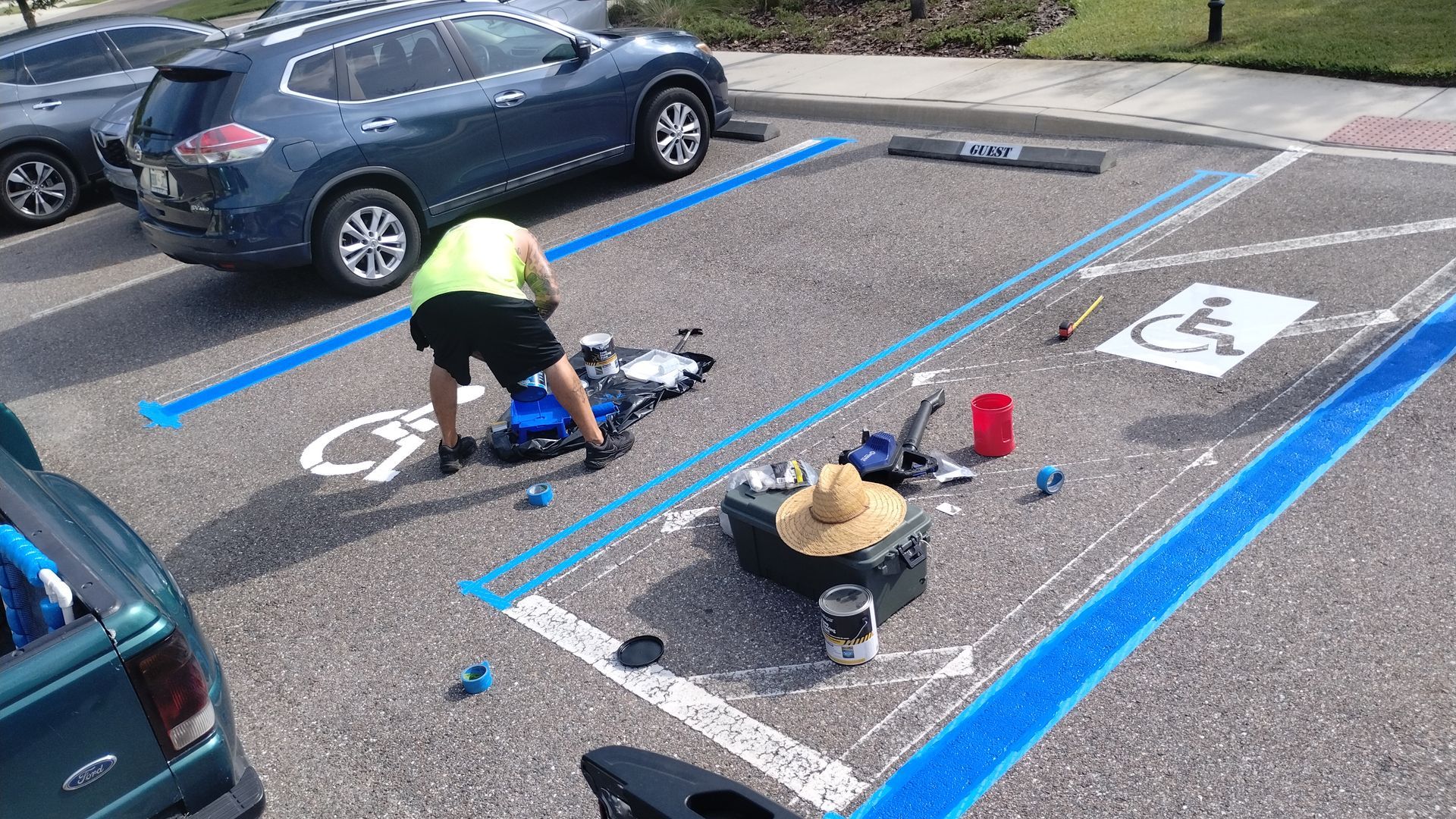 Man painting a blue handicapped parking space in a parking lot; tools and paint nearby; blue SUV in background.