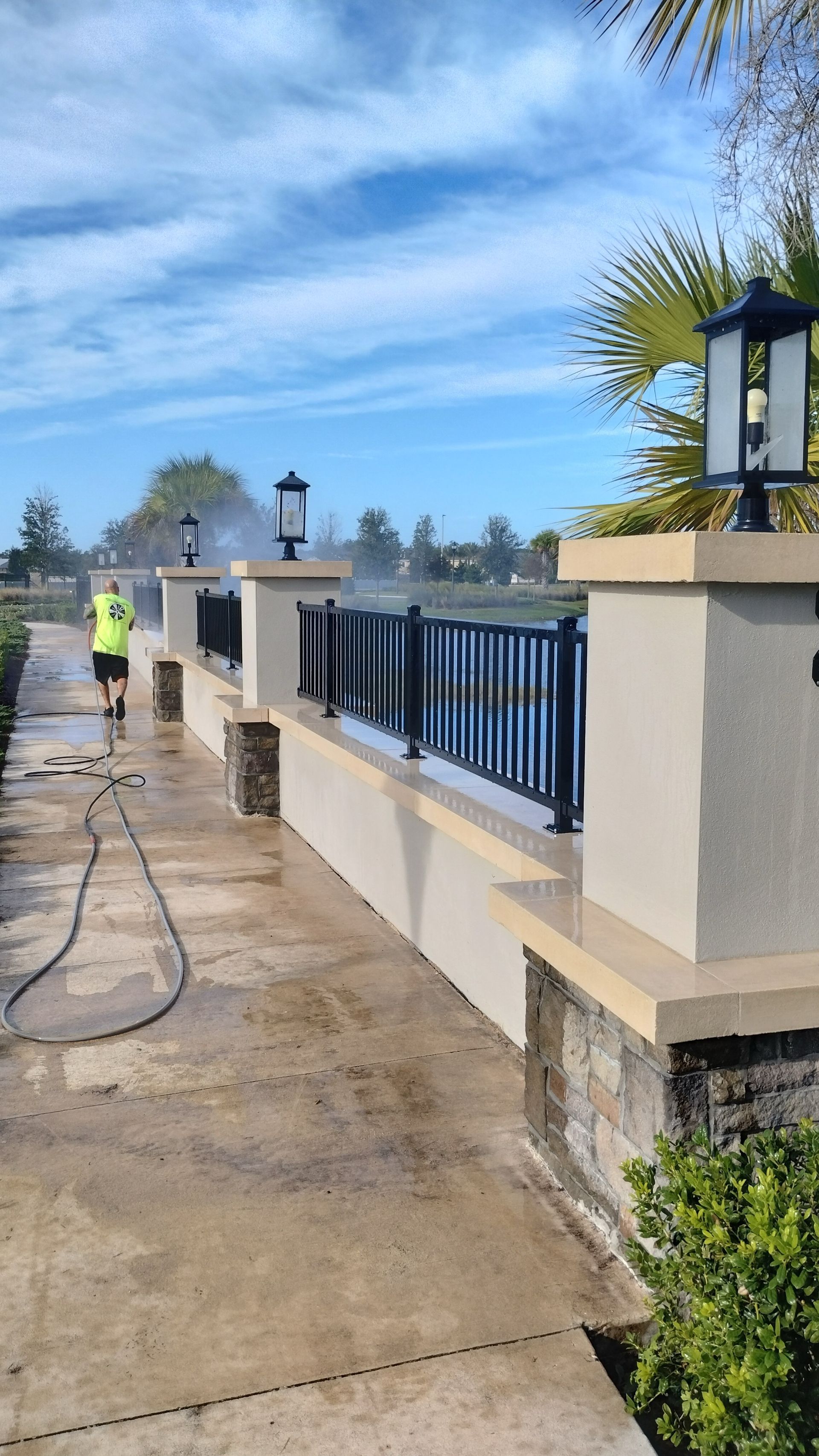 A person pressure washes a concrete walkway next to a low wall and black fence; sunny, blue sky.