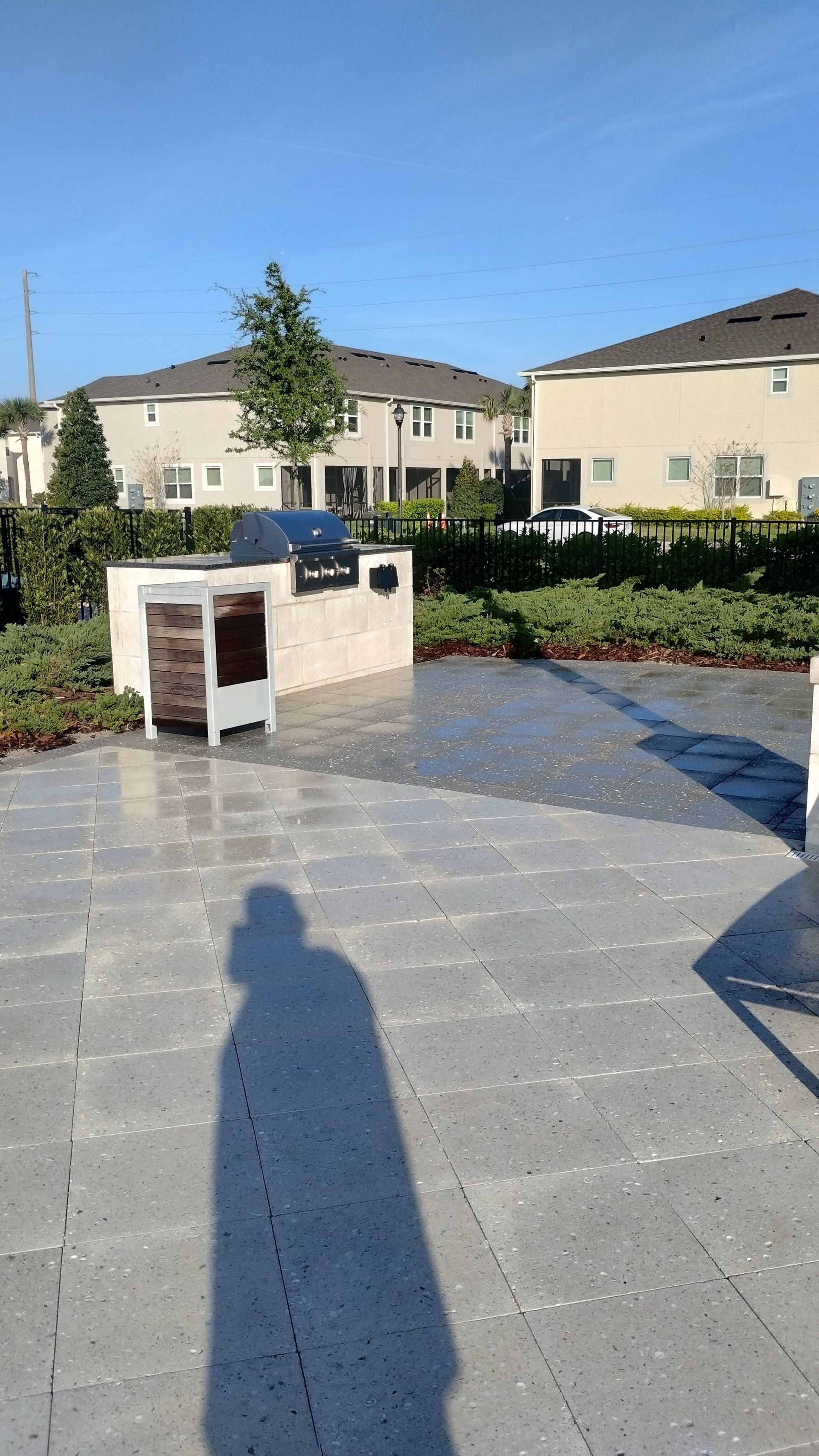 Patio with a built-in grill, person's shadow on the ground. Houses and bushes in the background, sunny day.