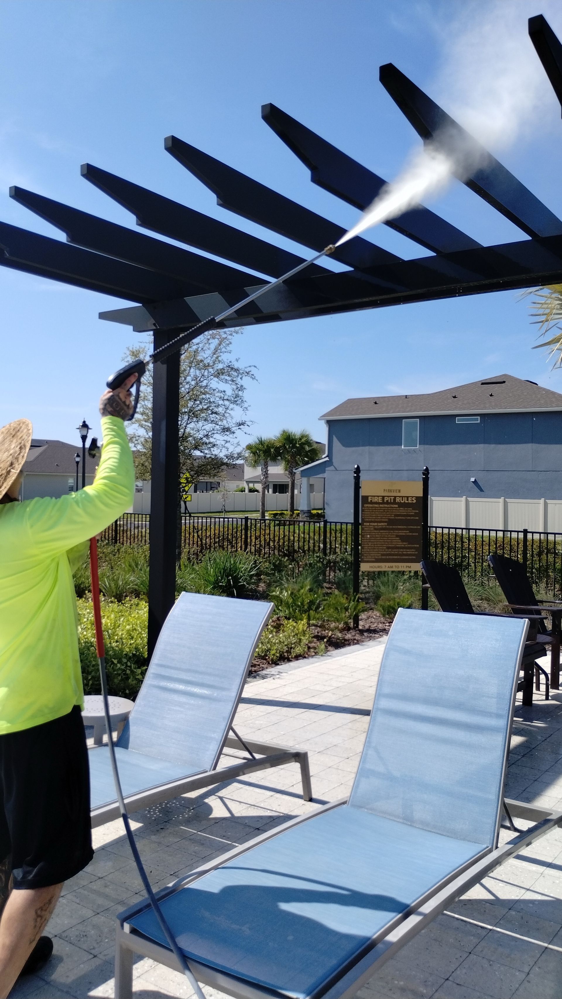 Person pressure washing a black pergola on a sunny day near two blue lounge chairs.