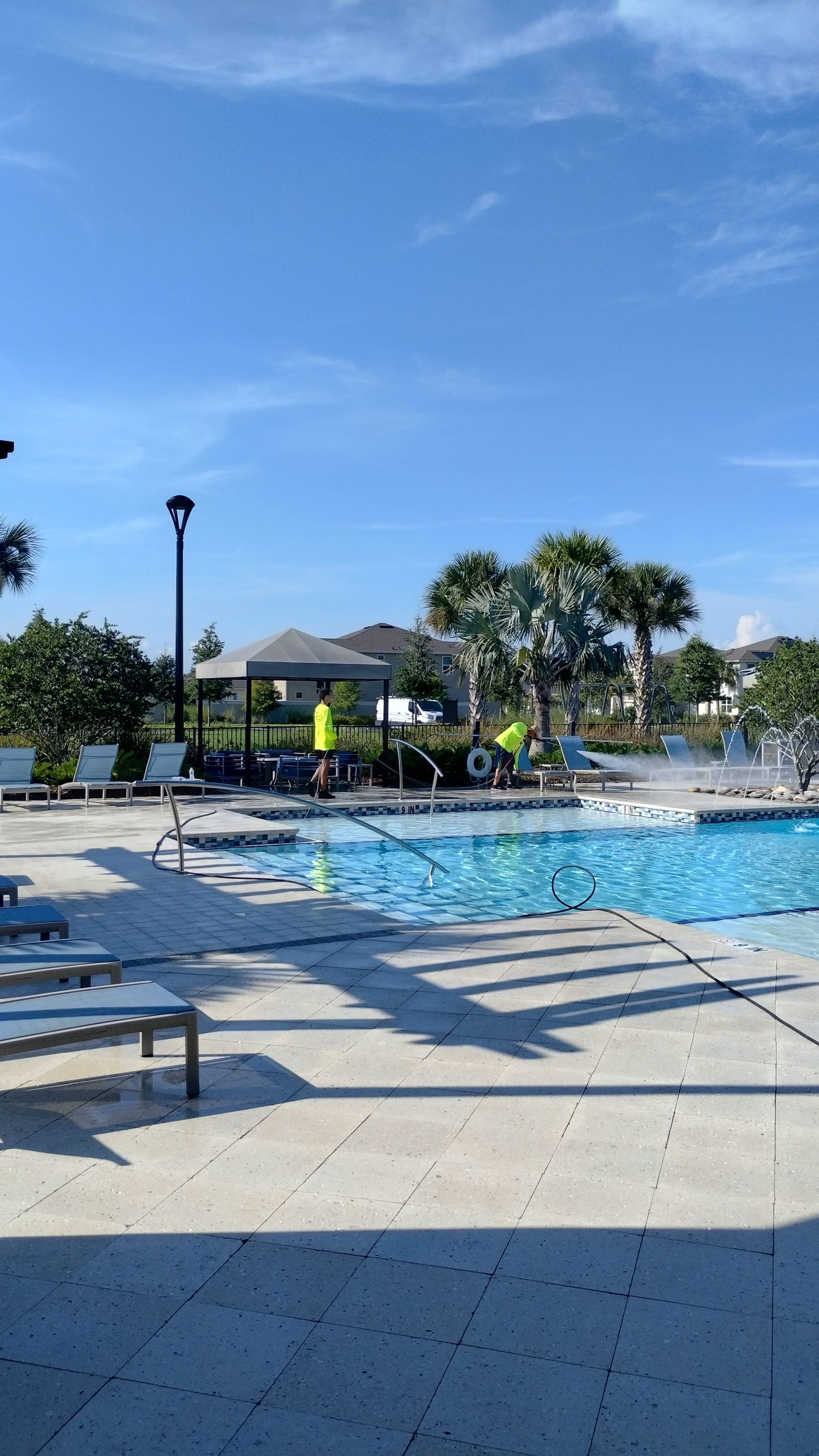 Pool area on a sunny day. Blue sky, clear pool, lounge chairs, trees, and a gazebo.