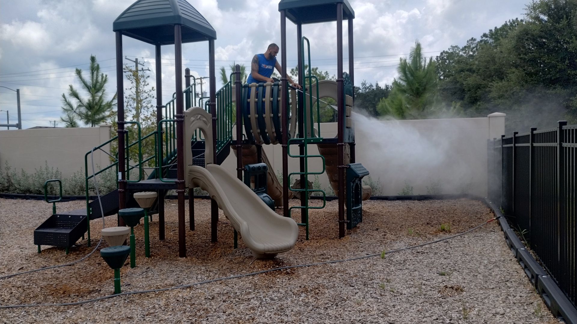 Man spraying a playground with mist. The play structure is green and tan, set on a bed of rocks next to a fence.