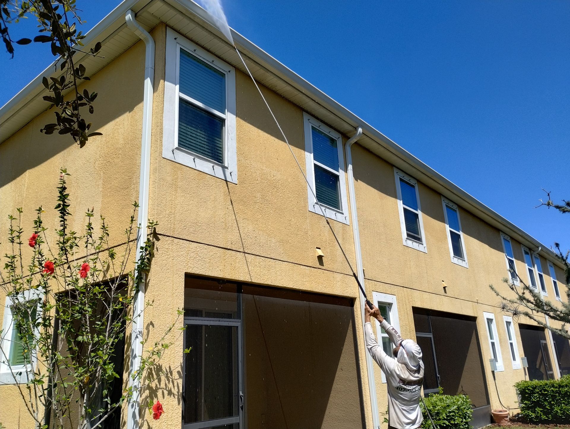Person washes a two-story yellow house with a long-handled sprayer on a sunny day.
