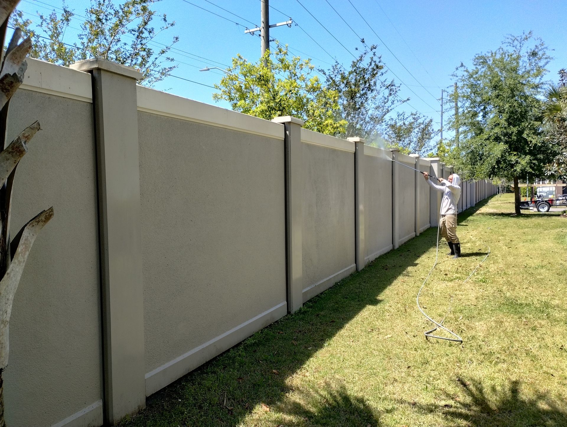 Man sprays tall beige concrete fence in a grassy yard under a bright blue sky.