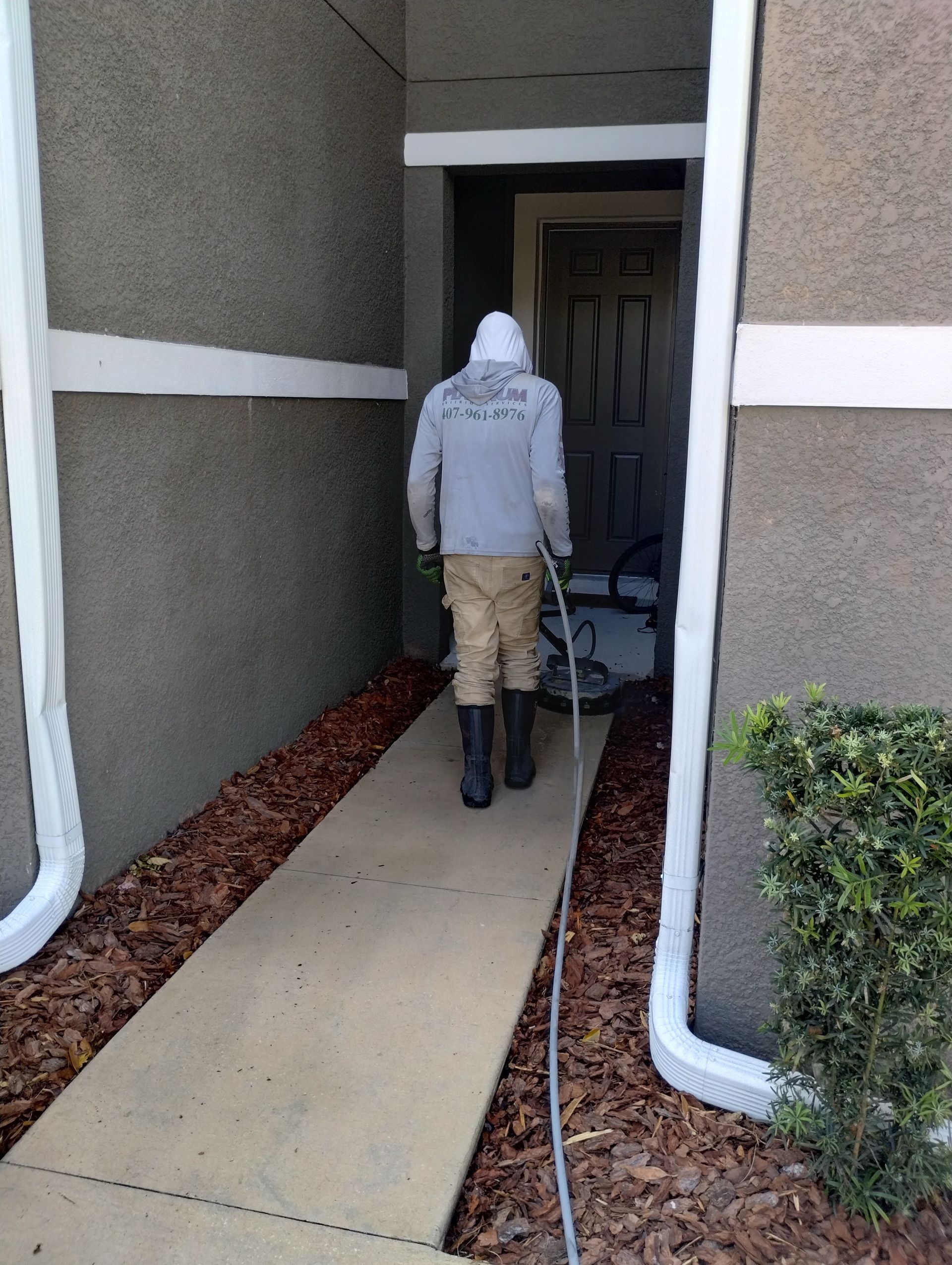 Person in protective gear walking toward a door, carrying a hose. Gray building exterior, pathway, mulch and greenery.
