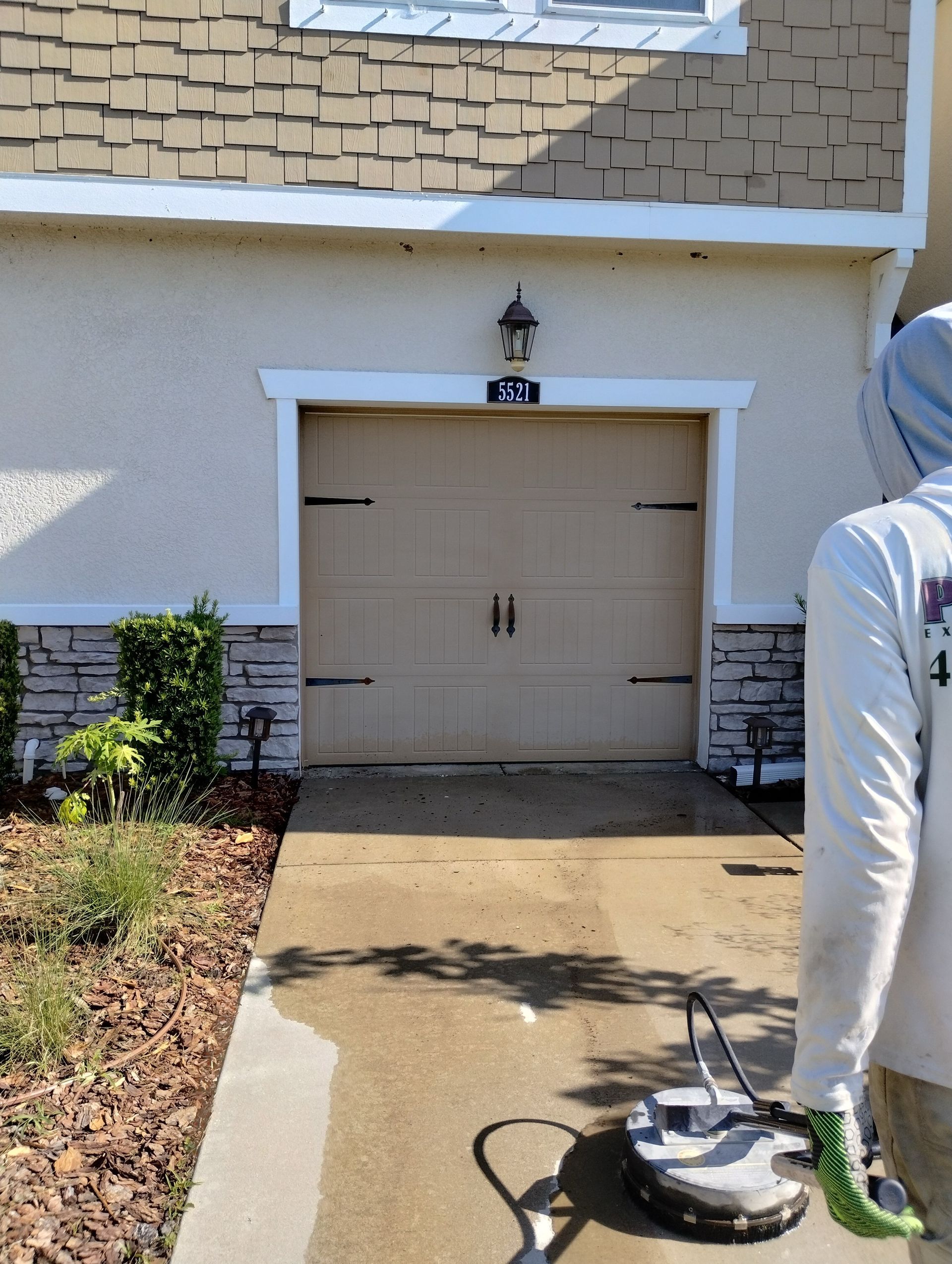 Person power washing a light brown garage door and driveway. Beige building with decorative stone and shrubbery.