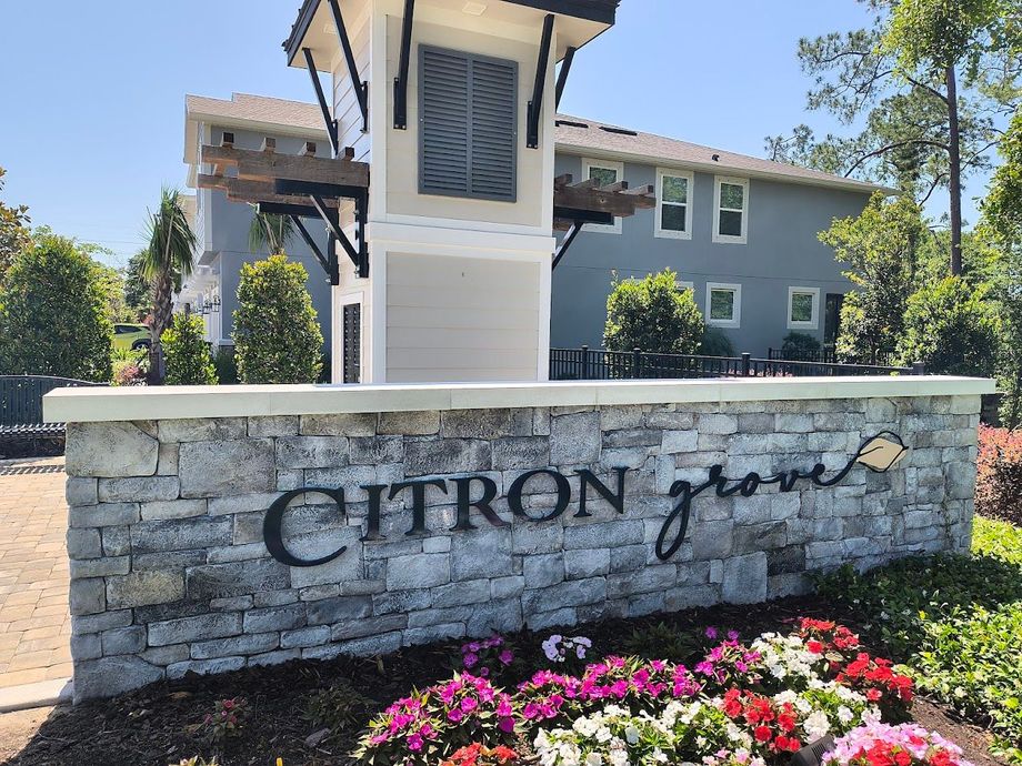 Stone sign for Citron Grove, with flowers in front and a two-story building in the background.