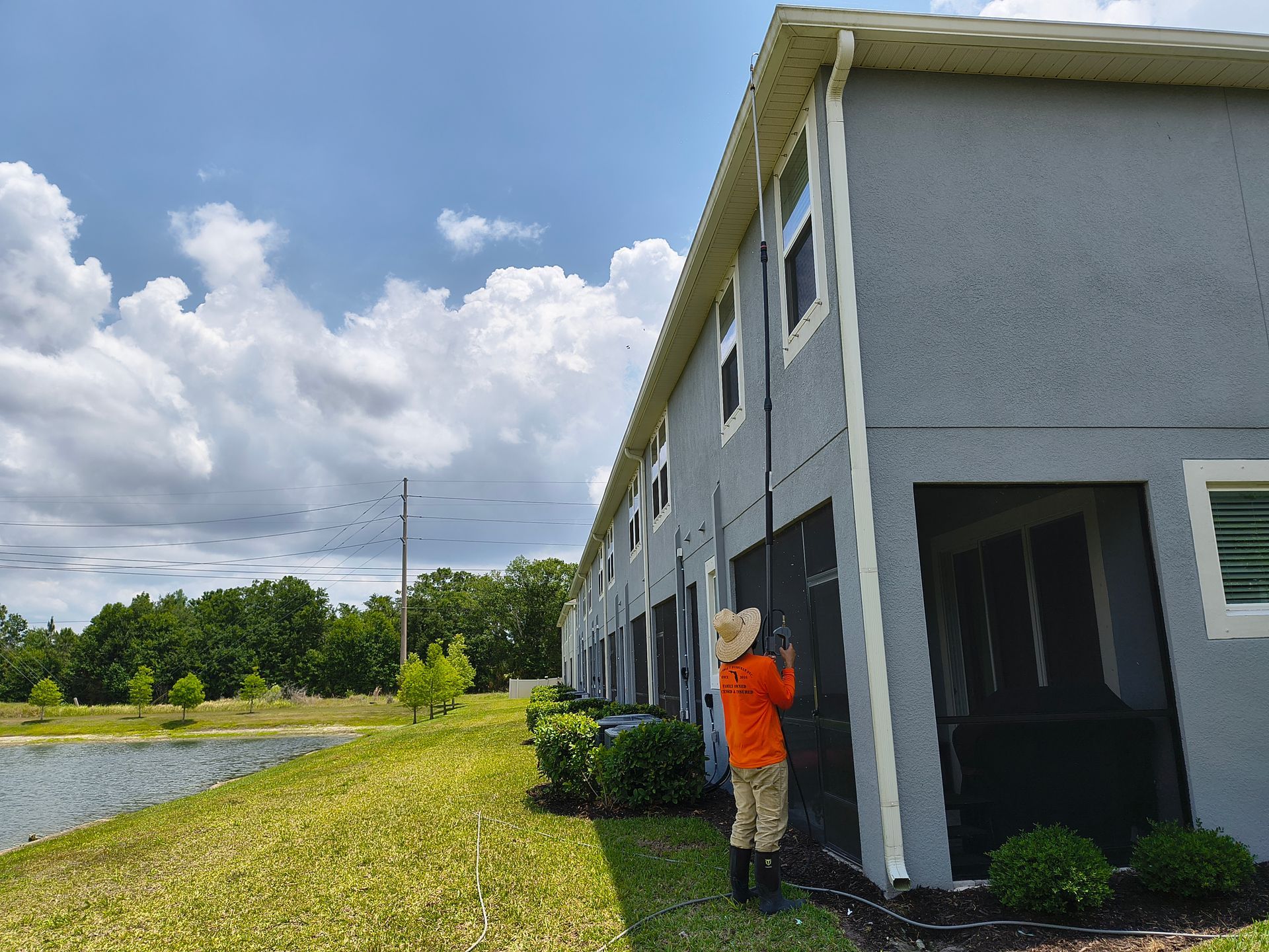 Person in orange shirt cleaning a grey building's side with a lake in the background under a cloudy sky.